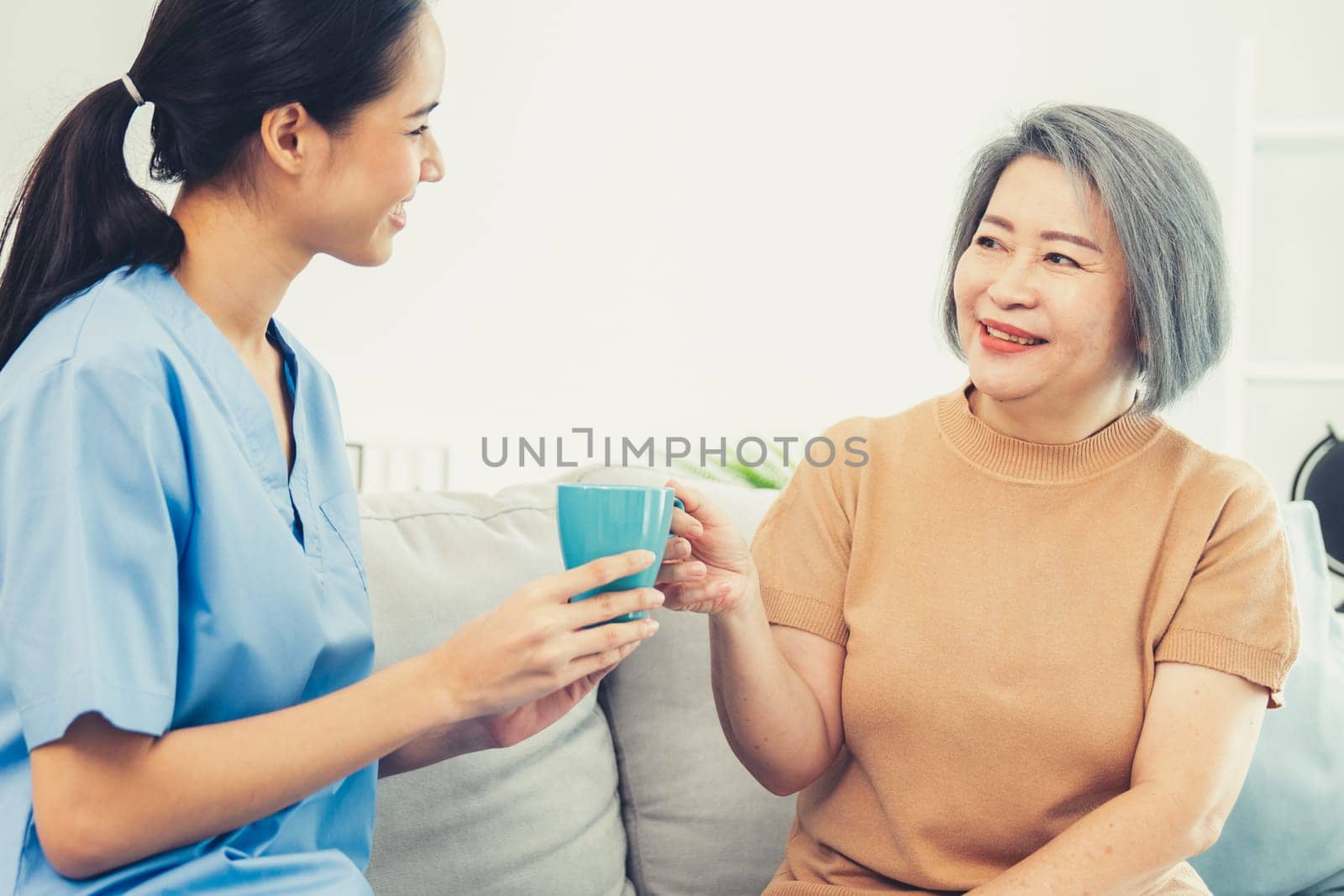 Female care taker serving her contented senior patient with a cup of coffee at home, smiling to each other. Medical care for pensioners, Home health care service.