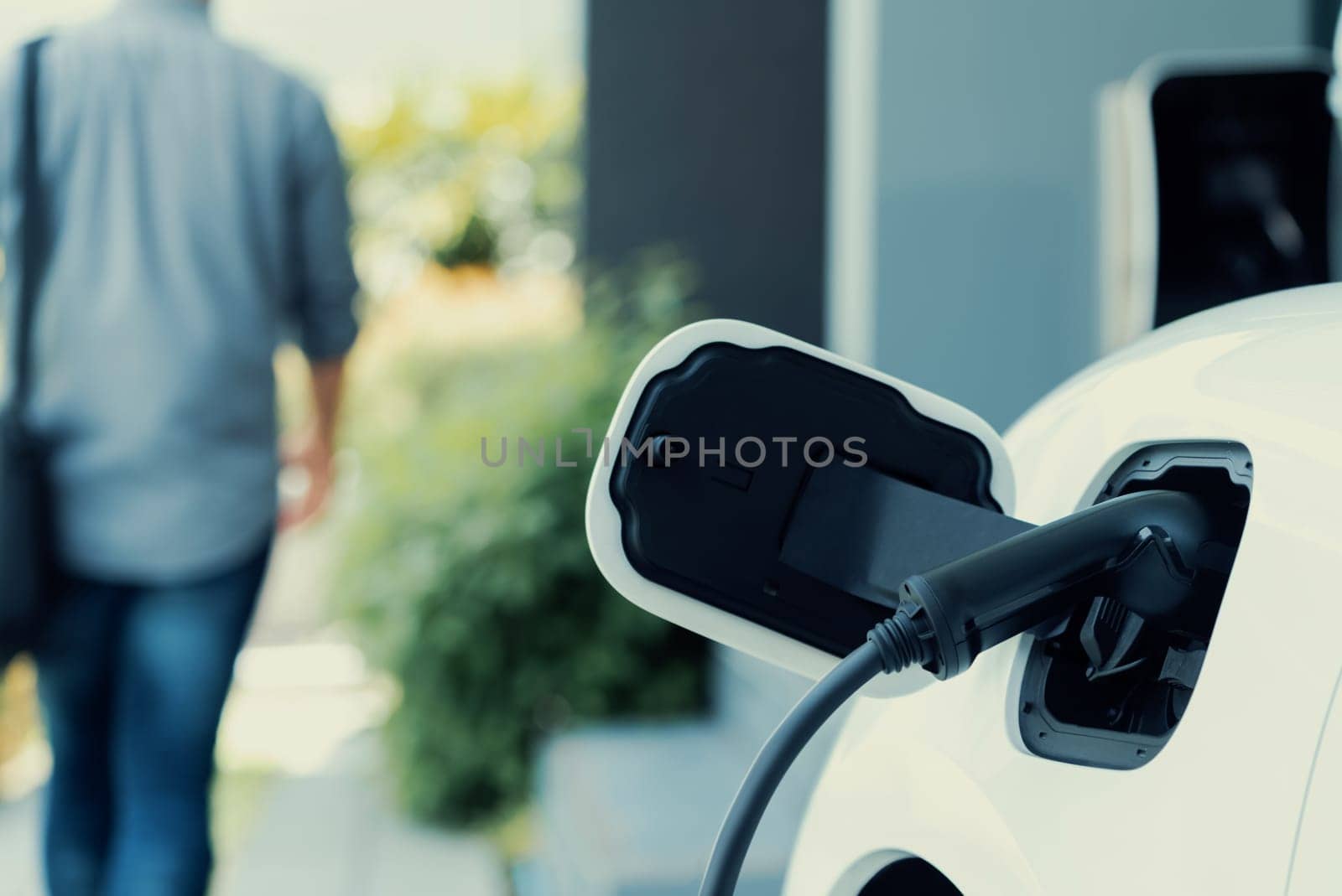 Focus electric car charging at home charging station with blurred progressive man walking in the background. Electric car using renewable clean for eco-friendly concept.