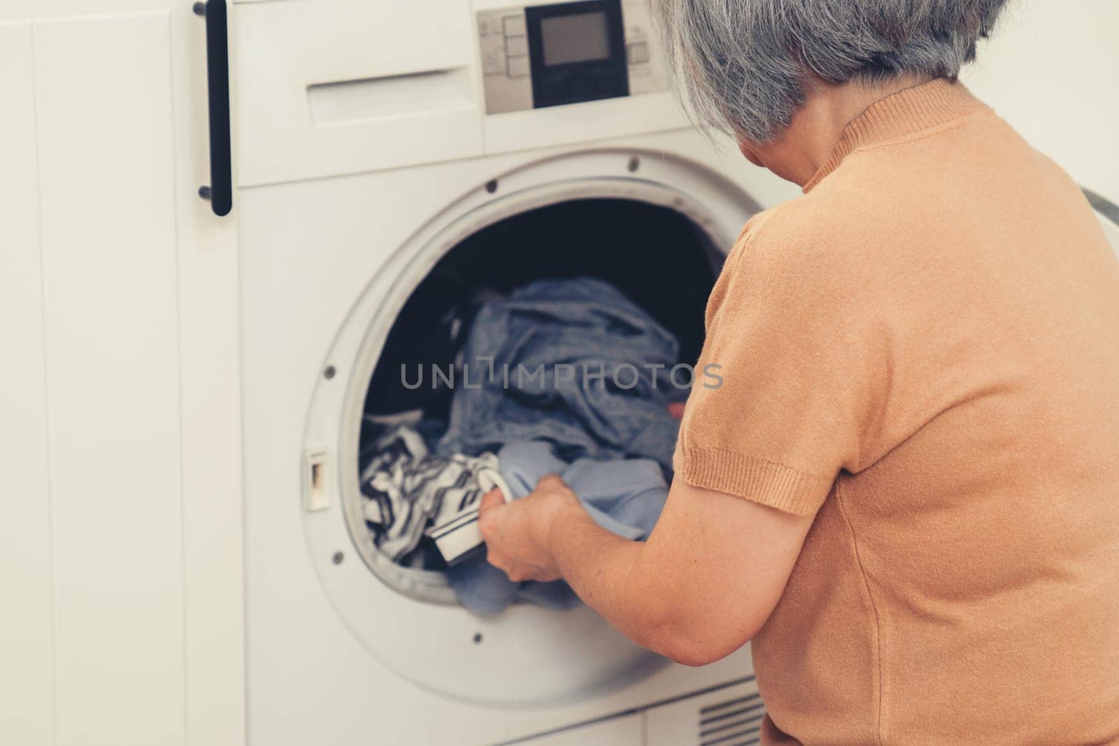 Contented senior housewife doing laundry in the laundry room with clothes inside the washing machine. Domestic life, drying machine, household chores.