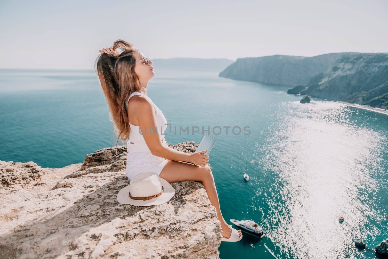 Successful business woman in yellow hat working on laptop by the sea. Pretty lady typing on computer at summer day outdoors. Freelance, travel and holidays concept.