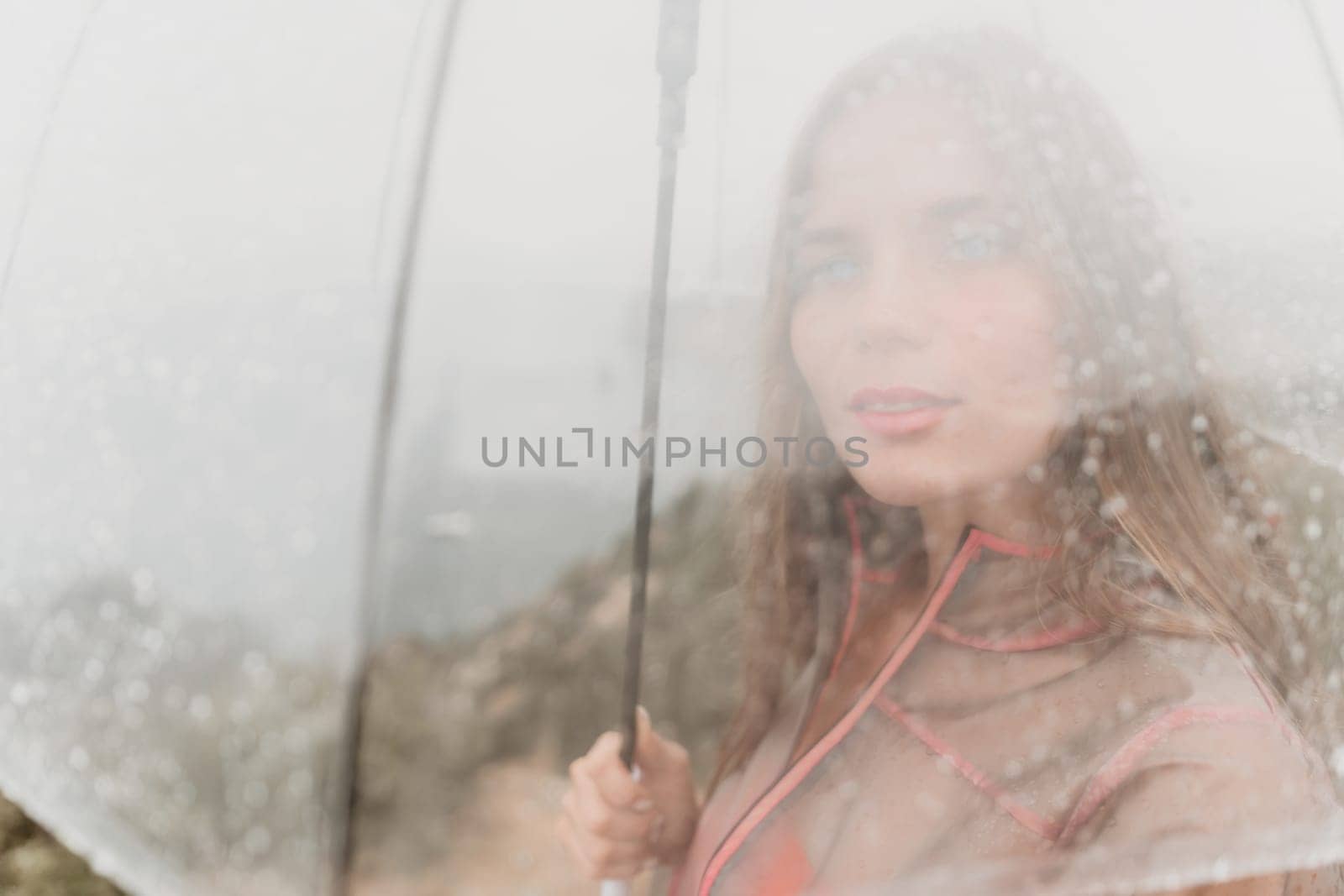 Woman rain umbrella. Happy woman portrait wearing a raincoat with transparent umbrella outdoors on rainy day in park near sea. Girl on the nature on rainy overcast day. by panophotograph