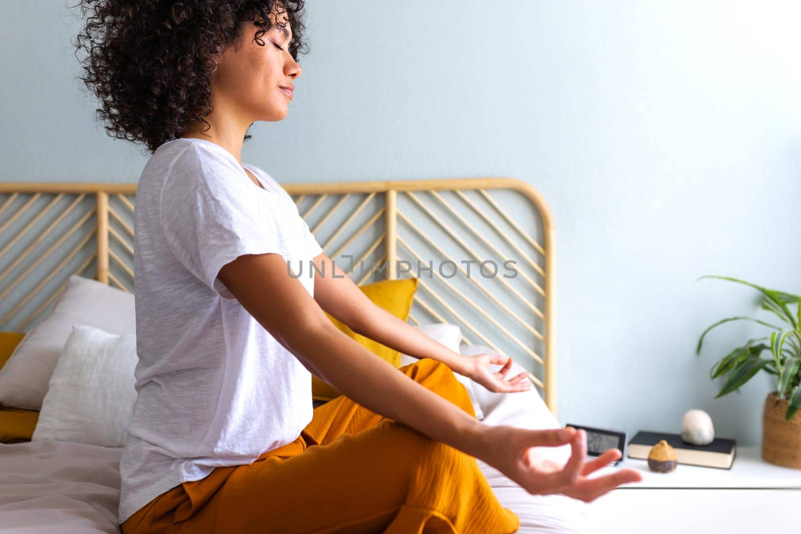 Side view of young multiracial woman relaxing on the bed. Meditating and breathing yoga exercises to calm mind. African American woman does meditation in bedroom. Wellness and meditation concept.