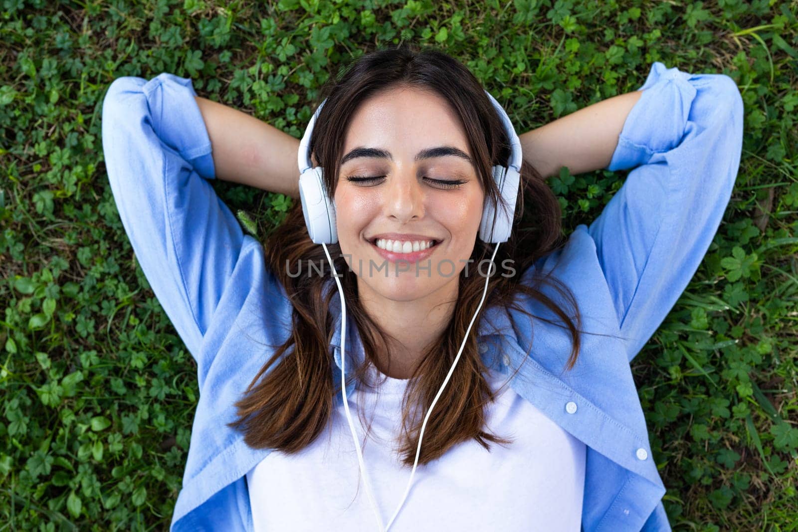 Top view of happy, smiling young caucasian woman relaxing lying down on grass with eyes closed listening to music with headphones. Wellbeing and lifestyle concept.
