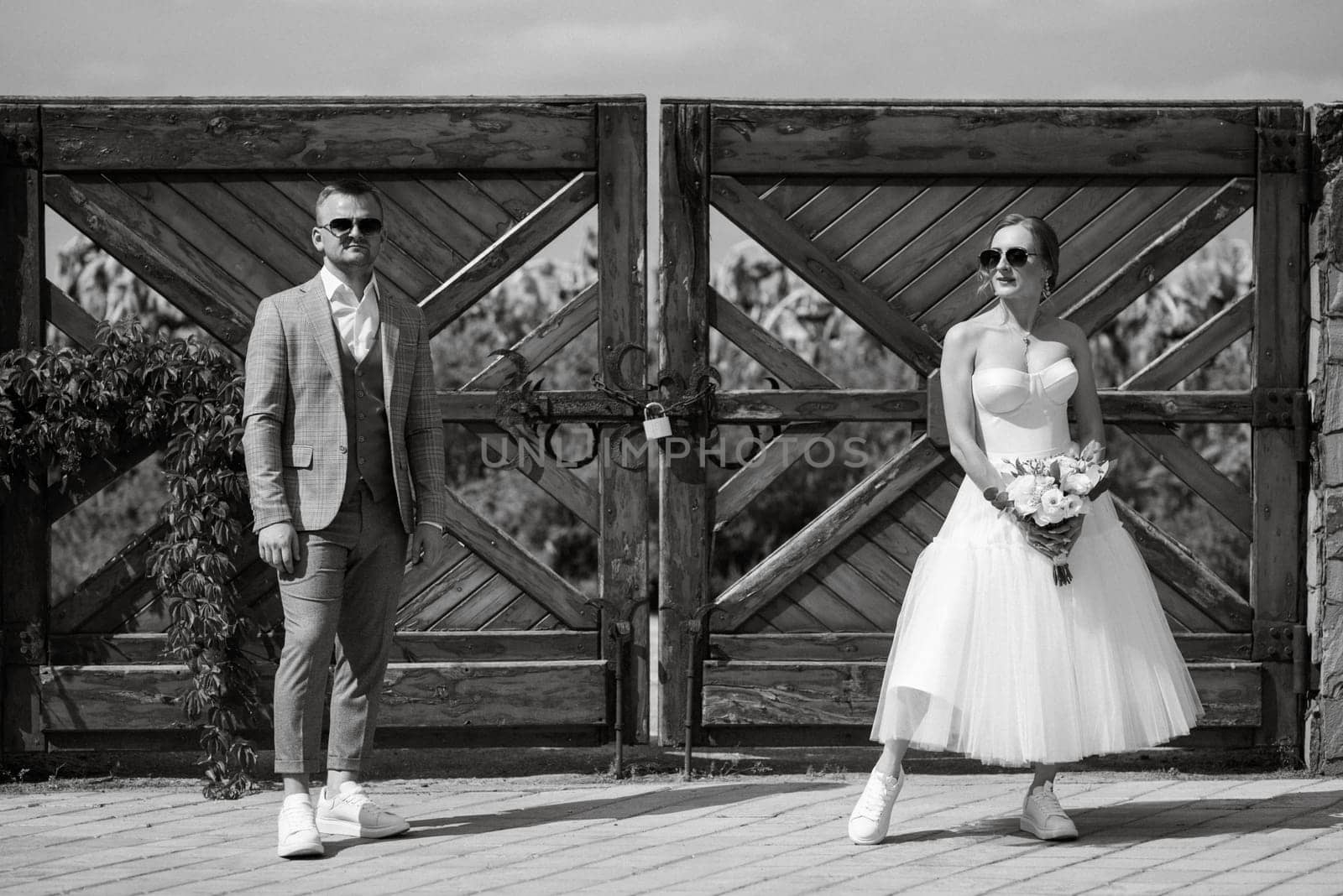 young couple bride in a white short dress and groom in a gray suit near the wooden rural gate of the estate