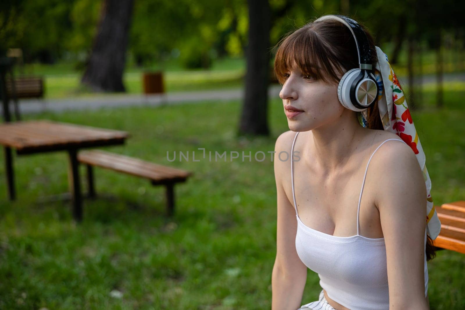 Woman in the park with wireless headphones on her head. Girl relaxes in the park listening to music. Relaxing in nature in summer. Woman in white strapless top.