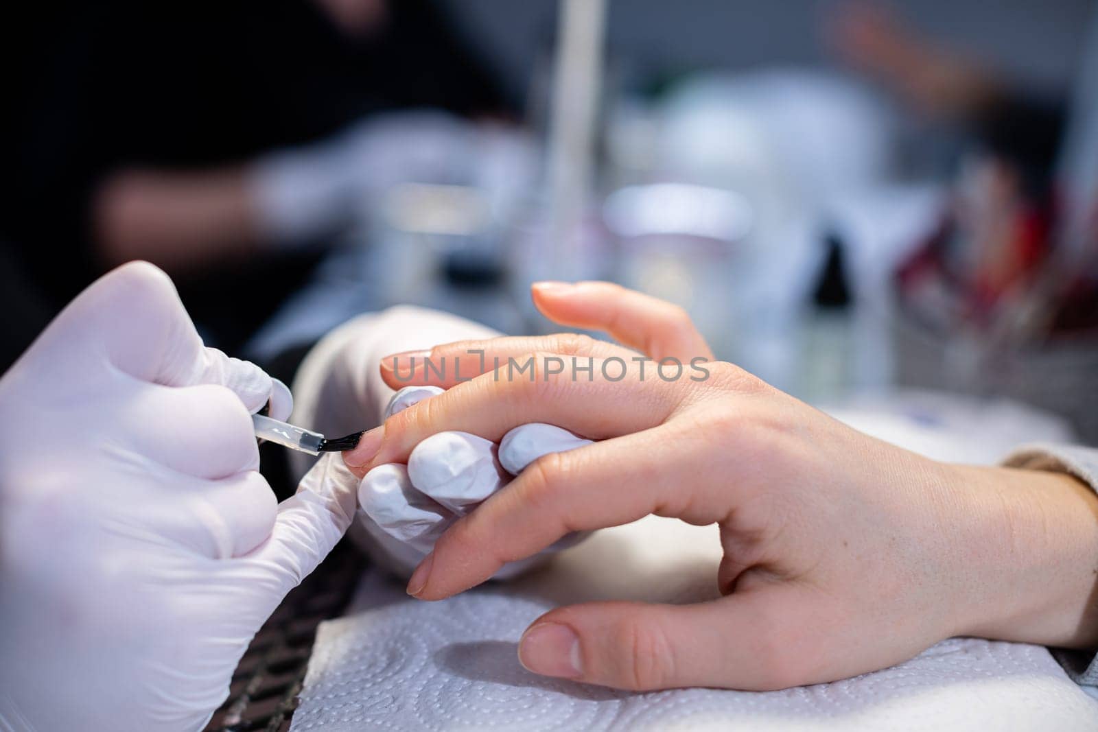 In the background the blurred interior of the beauty salon. The beautician wears disposable nitrile gloves. Performing a hybrid manicure on a natural nail plate. Work as a beautician and manicurist in a beauty salon.
