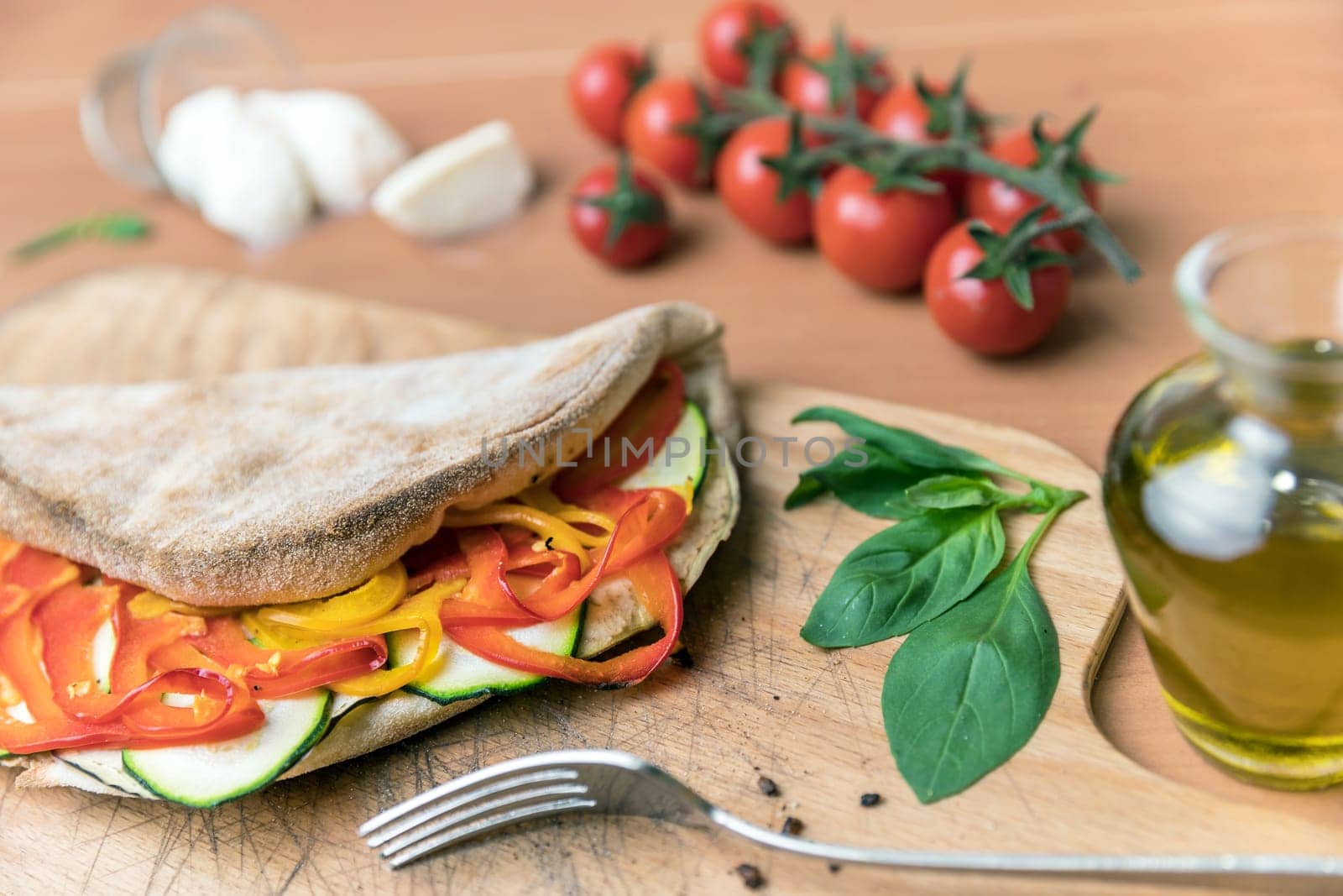 Italian food, closed vegetarian calzone pizza on rustic wooden background. Peppers and zucchini. On background small red tomatoes, mozzarella, basil and crude oil. Fork. Selective focus.
