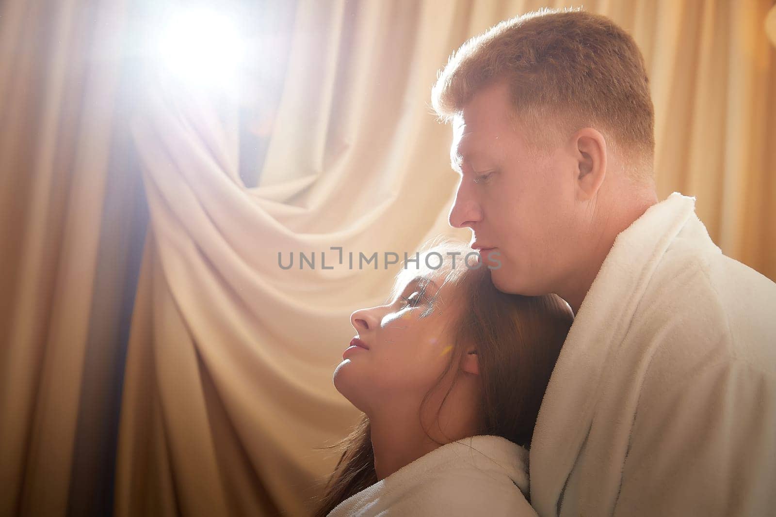 An adult couple of newlyweds in in white robes in a hotel room after the wedding. A guy and a girl, a man and a woman in beautiful room. The concept of hugs, love and care