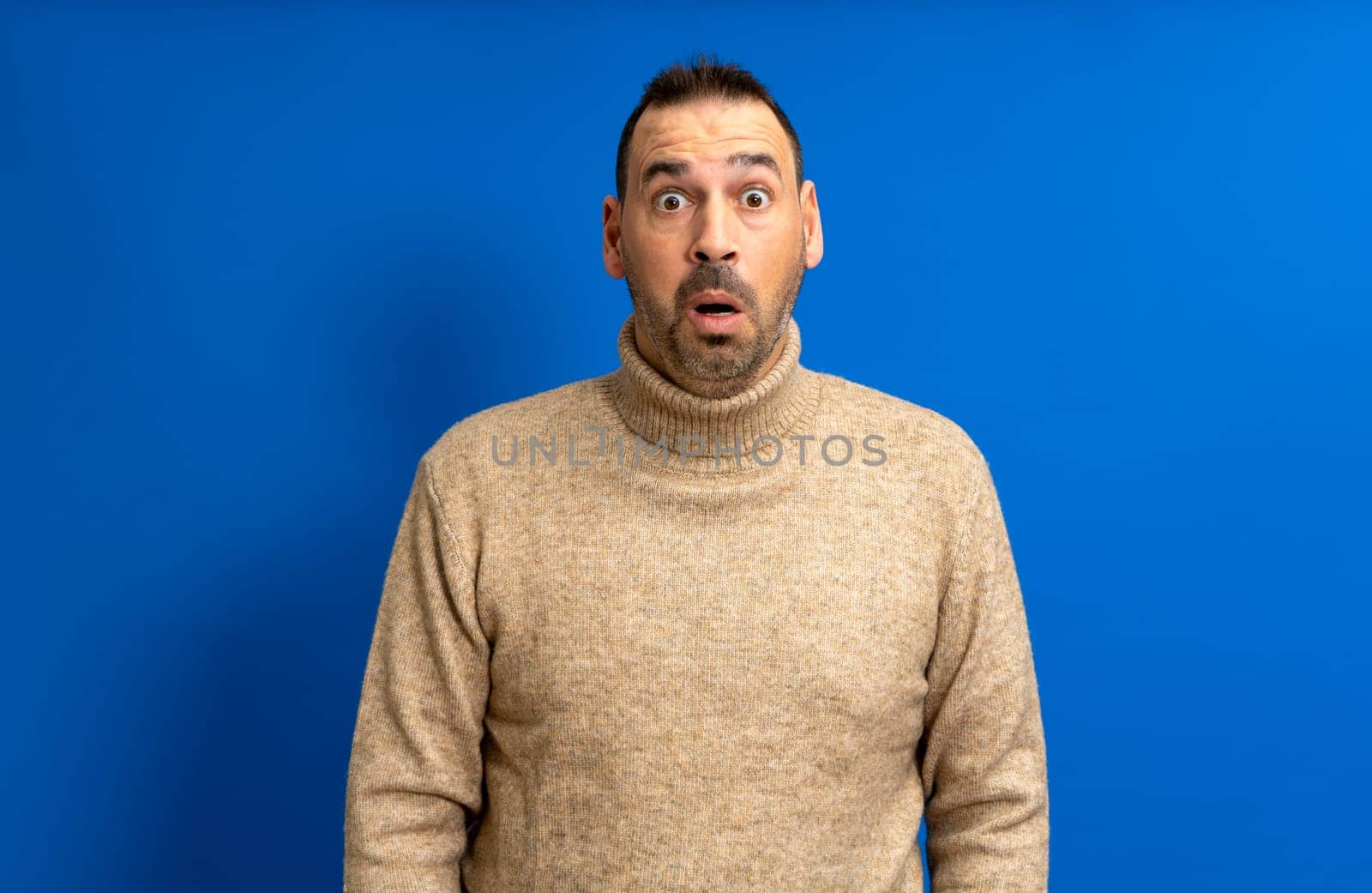 Bearded hispanic man wearing a turtleneck sweater with open mouth amazed and absorbed in something isolated over blue studio background