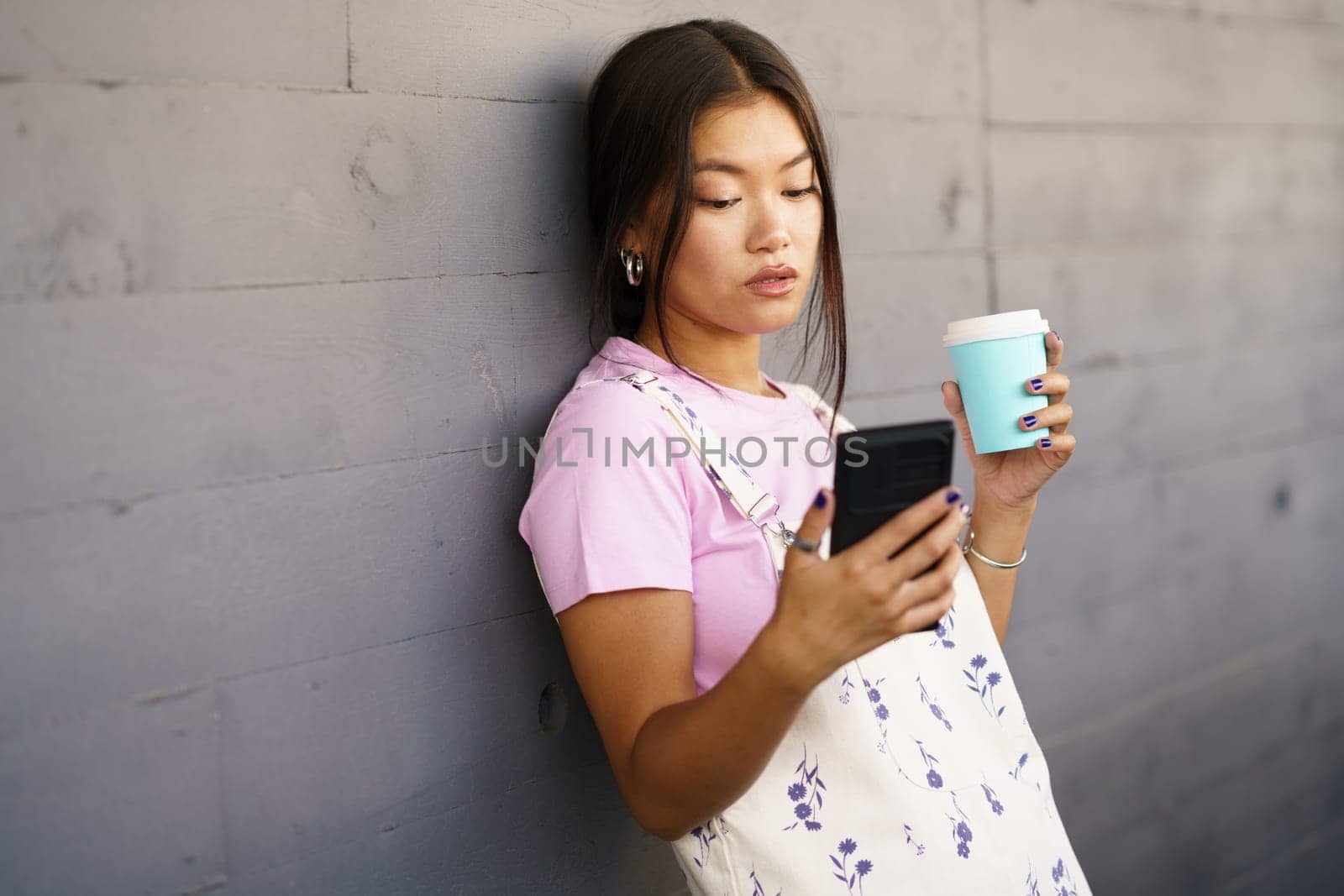 Asian female in stylish outfit with takeaway cup of coffee leaning on wall and reading news on cellphone in daytime on city street