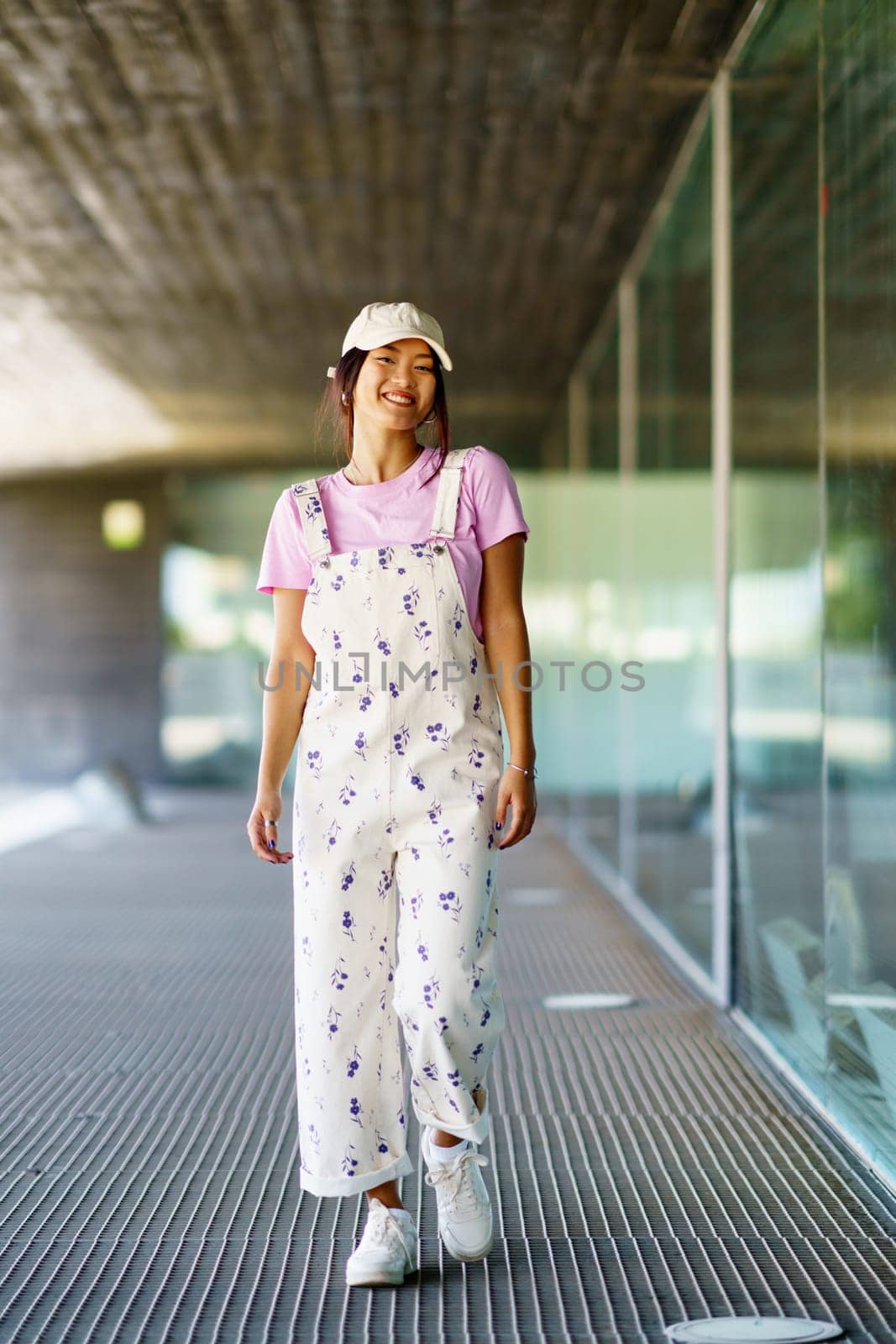 Full body happy young Asian female in stylish clothes and cap, smiling and looking at camera while walking near glass wall of contemporary building in daytime