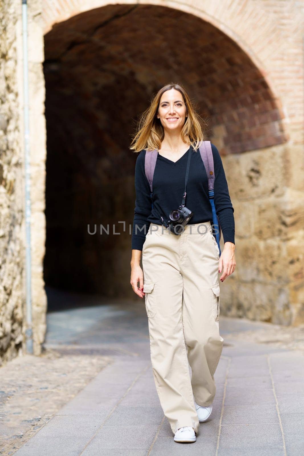 Full body of positive young female traveler with long hair in casual clothes and backpack, with photo camera hanging on neck smiling while walking in old city near arched passage