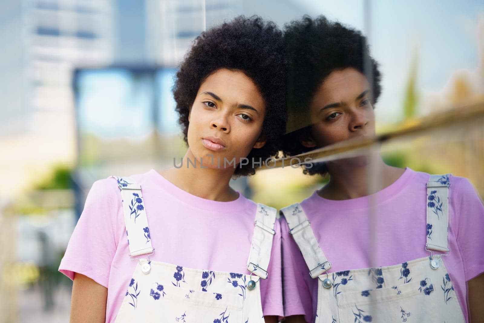 Serious young African American female with Afro hairstyle looking at camera while standing near glass house on street against blurred background