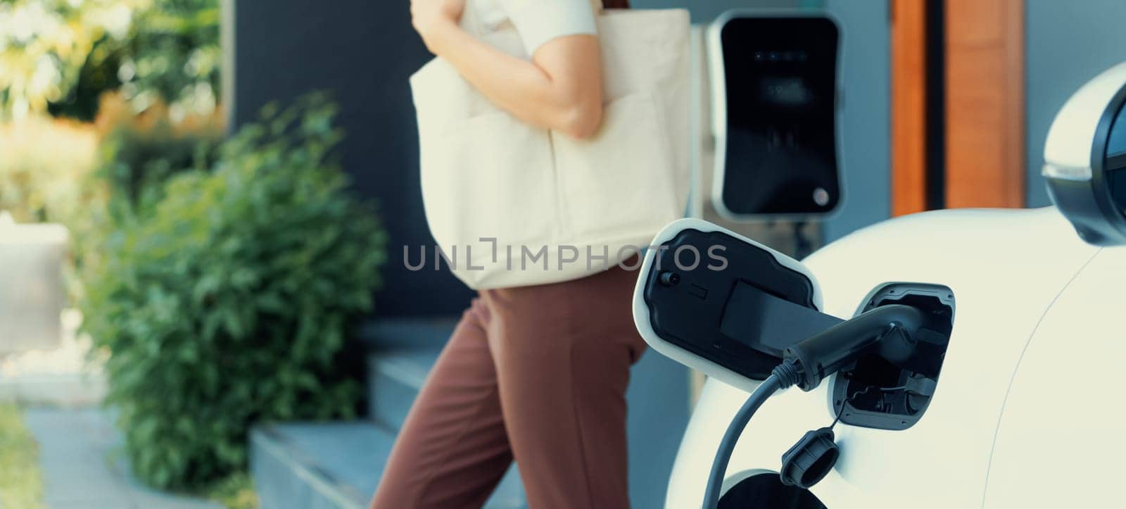 Focus image of electric vehicle recharging battery at home charging station with blurred woman walking in the background. Progressive concept of green energy technology applied in daily lifestyle.