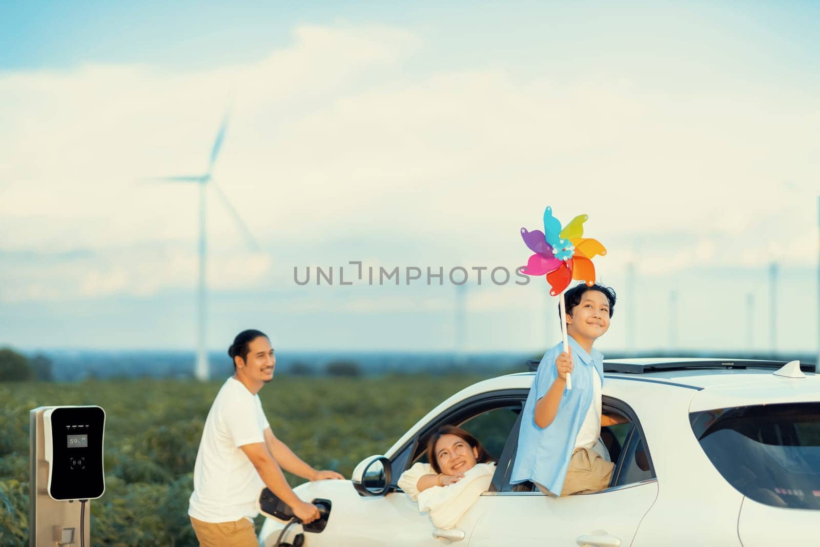 Concept of progressive happy family enjoying their time at wind farm with electric vehicle. Electric vehicle driven by clean renewable energy from wind turbine generator for charging station.