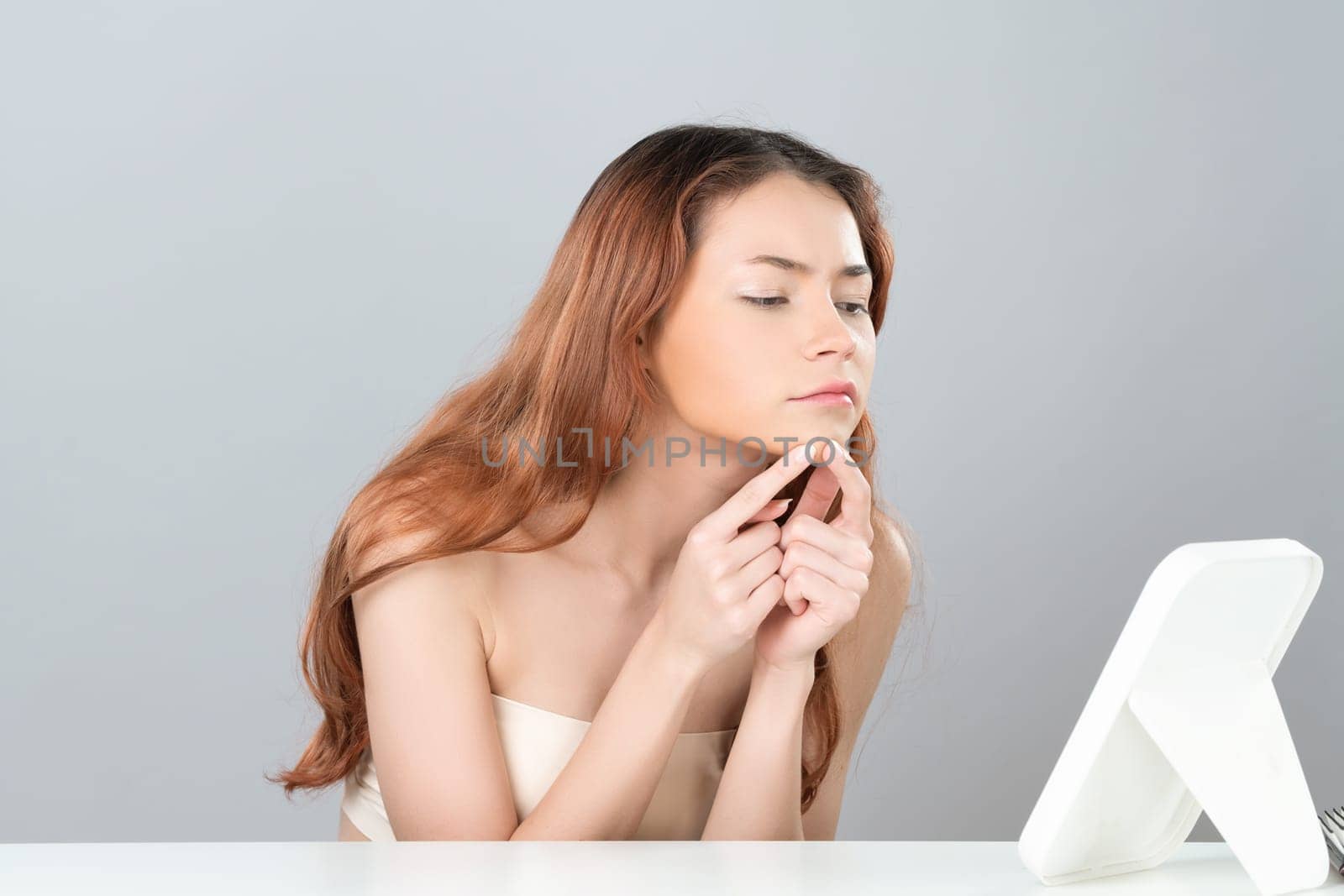 Glamorous portrait of beautiful woman feel insecure on acne spots problem. Worried female model with perfect smooth clean skin checking her face on mirror in isolated background.