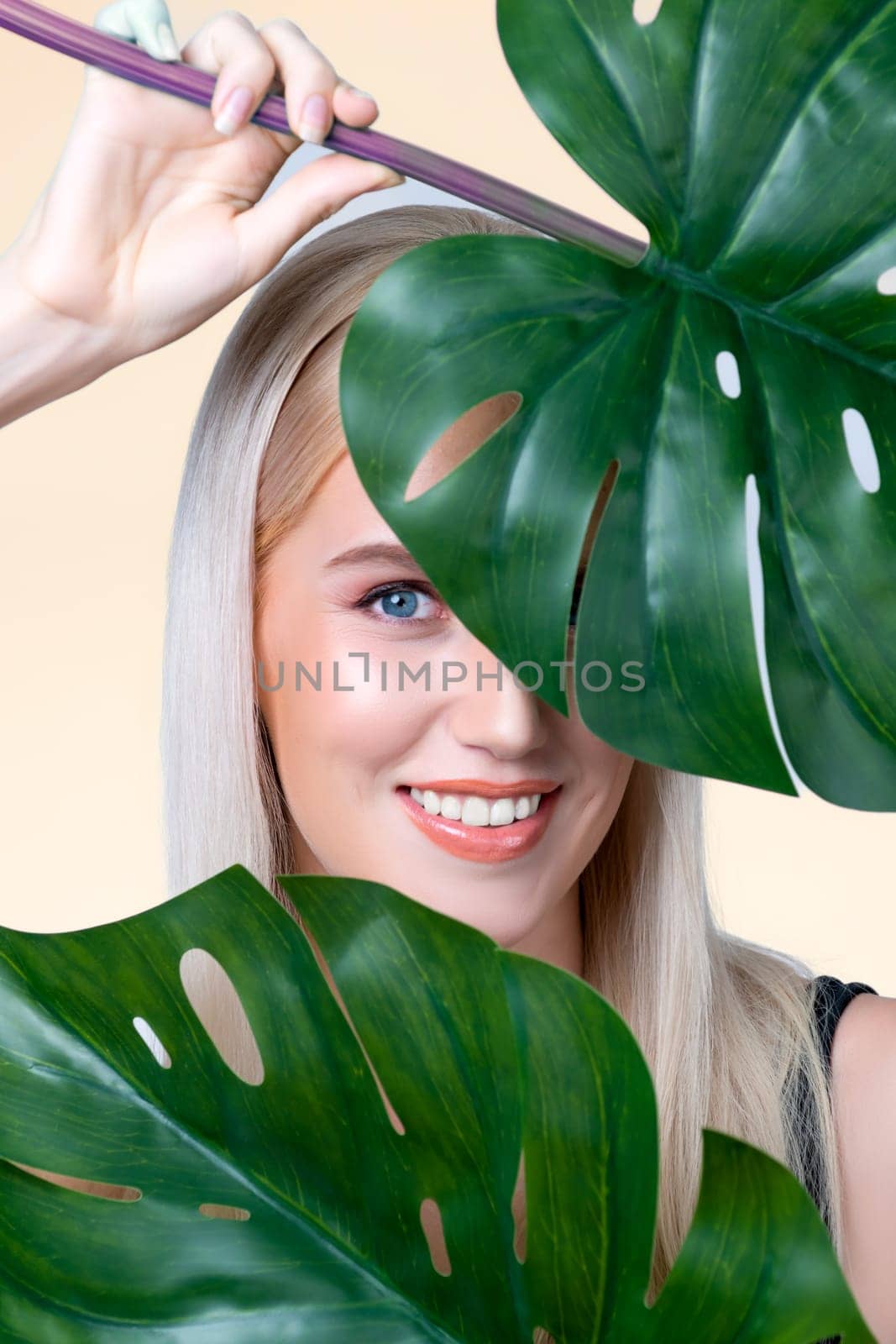 Closeup facial portrait personable woman with perfect smooth makeup holding green monstera leaves and cover her face as natural healthy skincare treatment. Tropical nature and beauty concept