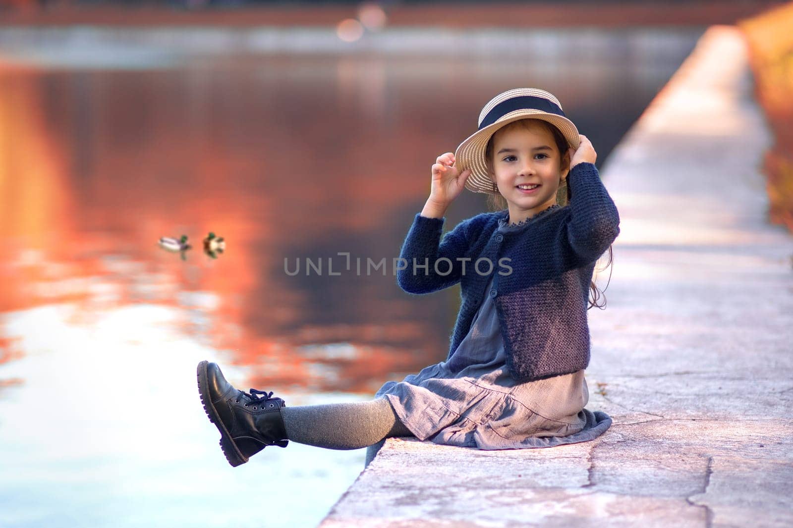 A cheerful little girl sits on the shore of a pond, smiling in vintage clothes, a straw hat, a gray dress and a knitted blouse against a background of a park and red foliage in autumn. Copy space