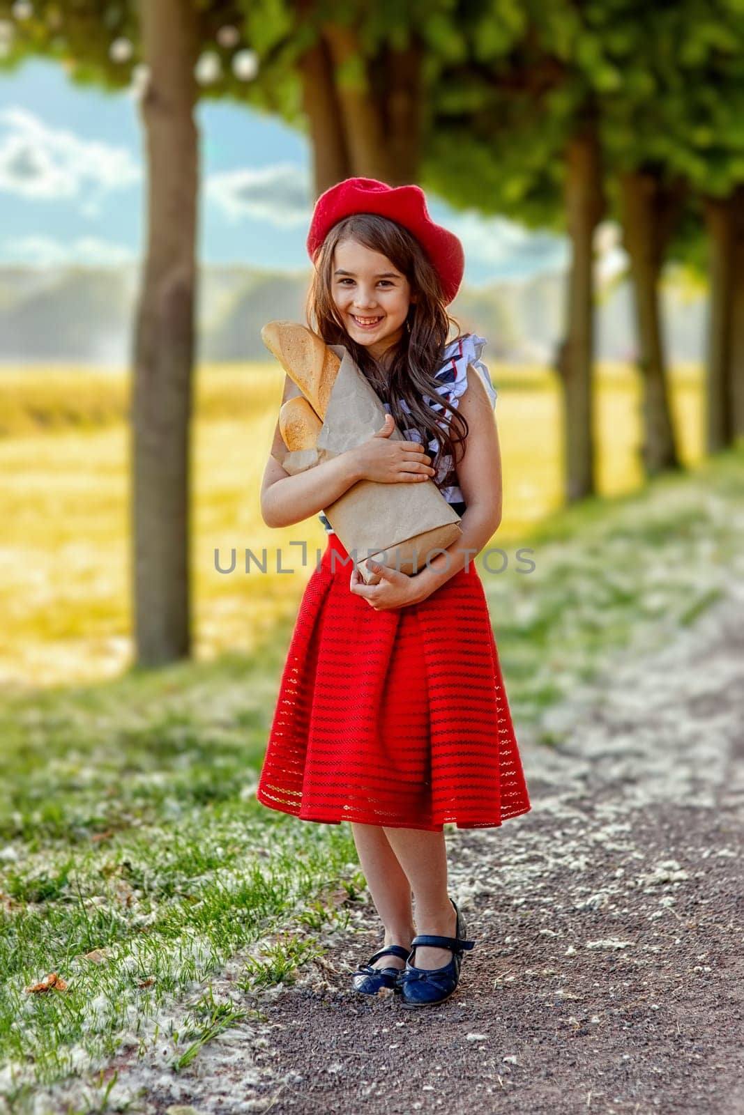 A beautiful brunette little girl in a red beret and a striped T-shirt stands against the background of green trees in the park, holding a craft bag with French baguettes. Copy space