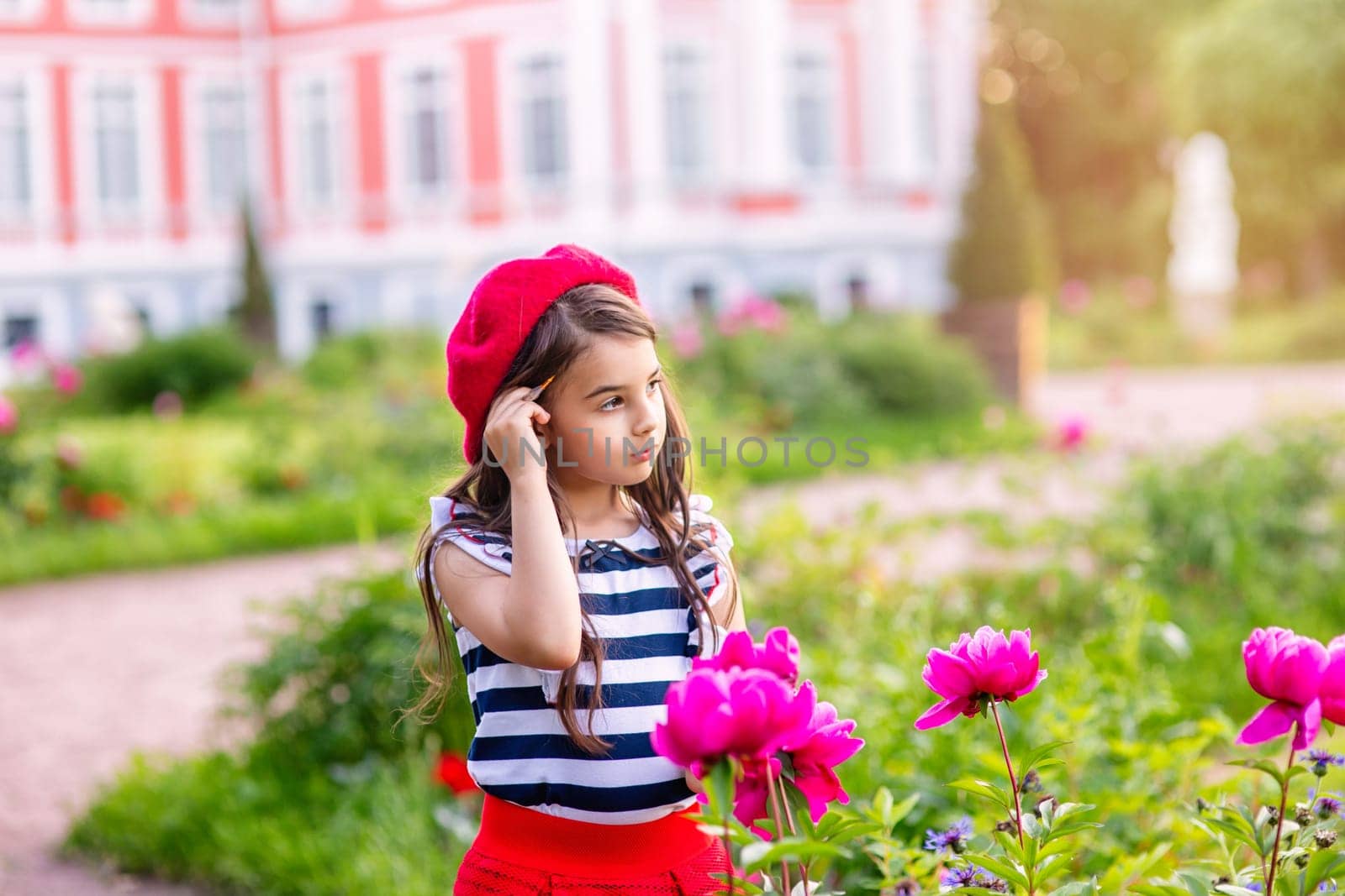 A beautiful little girl, an artist, in a red beret and a striped T-shirt stands next to a bush of peonies in a park in summer at sunset. Copy space