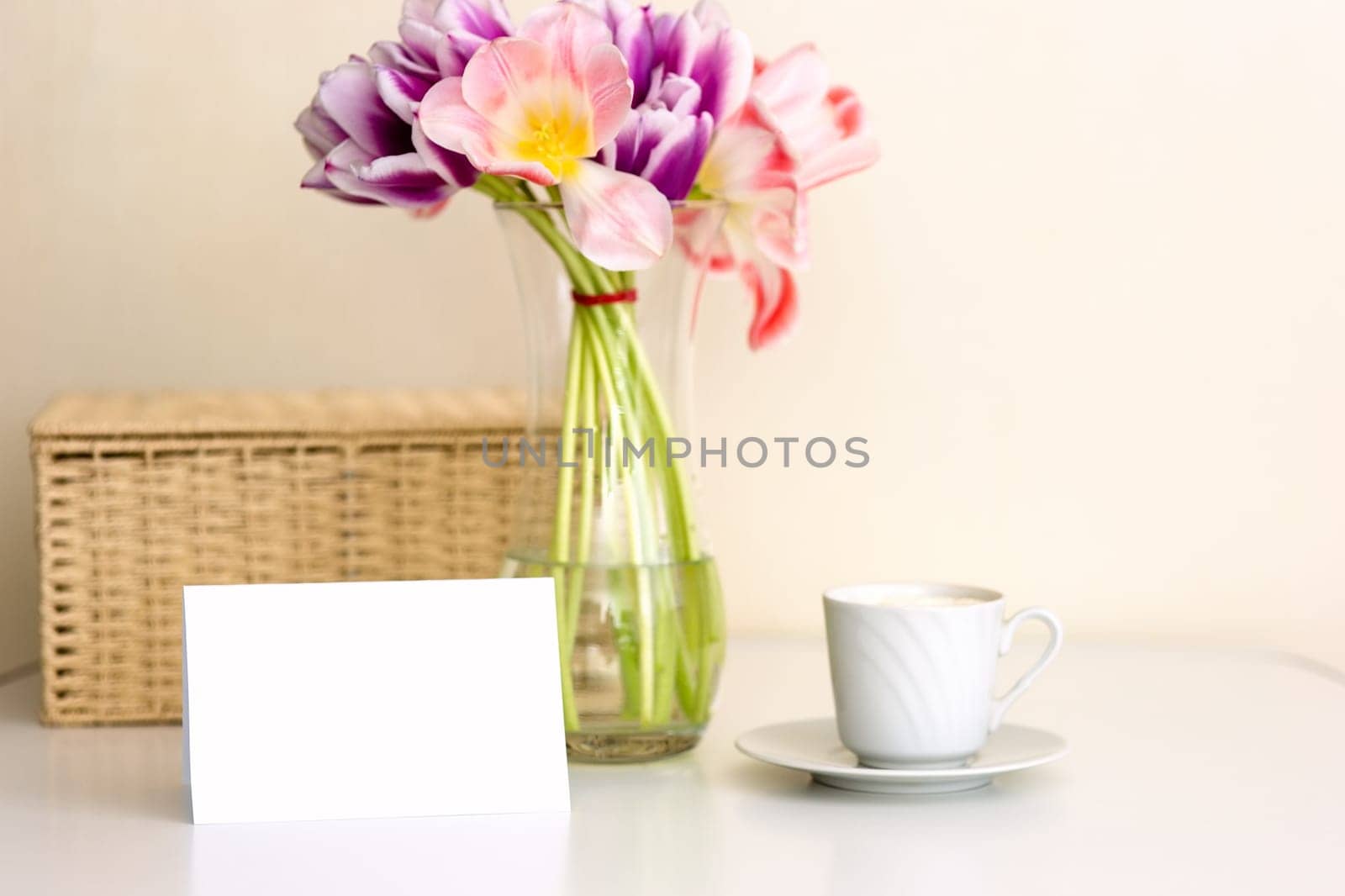 A bouquet of opened pink and purple tulips, a wicker basket, a white cup and an empty postcard with space for text, on a white table in a bright room. Cozy home concept