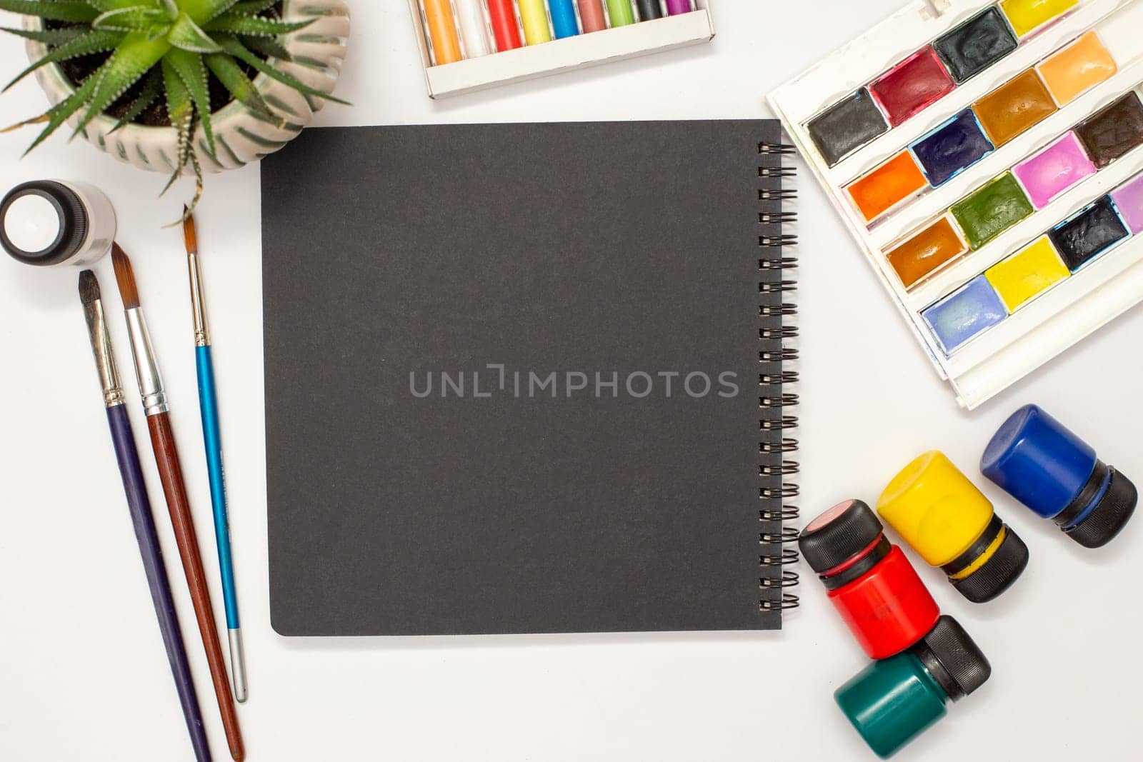 Black square sketchbook on a white table with drawing supplies: watercolors, pastel crayons, brushes, acrylic paints, succulent pot.Copy space.Mock up