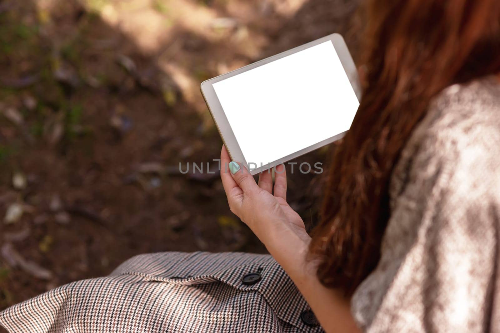 A woman with long brown hair, sitting in a knitted jumper in the park, holds a digital tablet. Remote work, education concept. Copy space. No face. Mock up