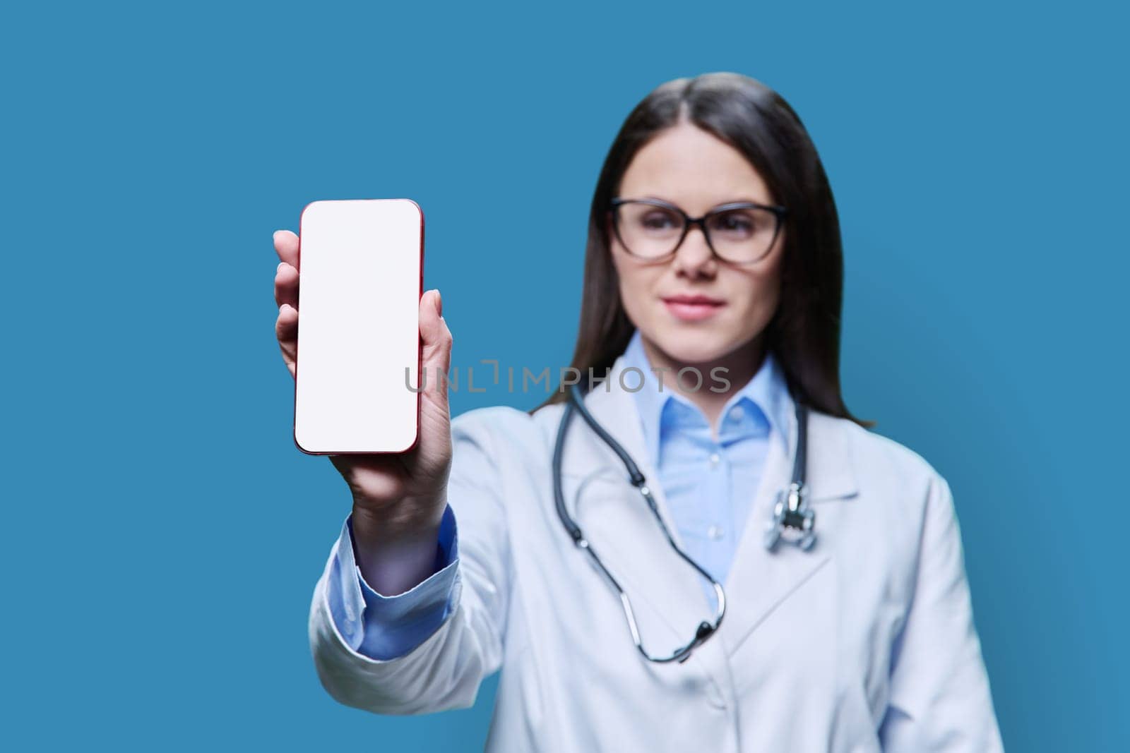Close-up of a blank screen smartphone in the hands of a female doctor on a blue studio background. Mobile apps, ads, medicine technology concept