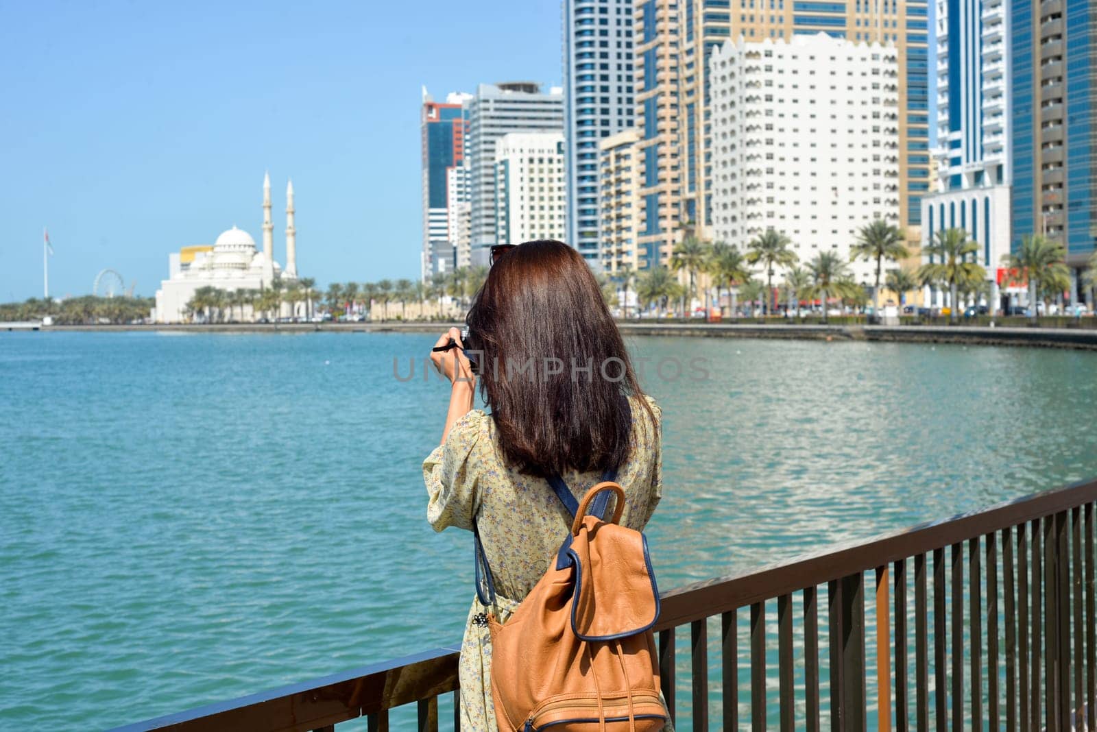 A woman in a long dress with a backpack walks with a camera along the Al Majaz embankment, Lake Khaled, Sharjah emirate. Rear view of a woman photographing the embankment by Ekaterina34