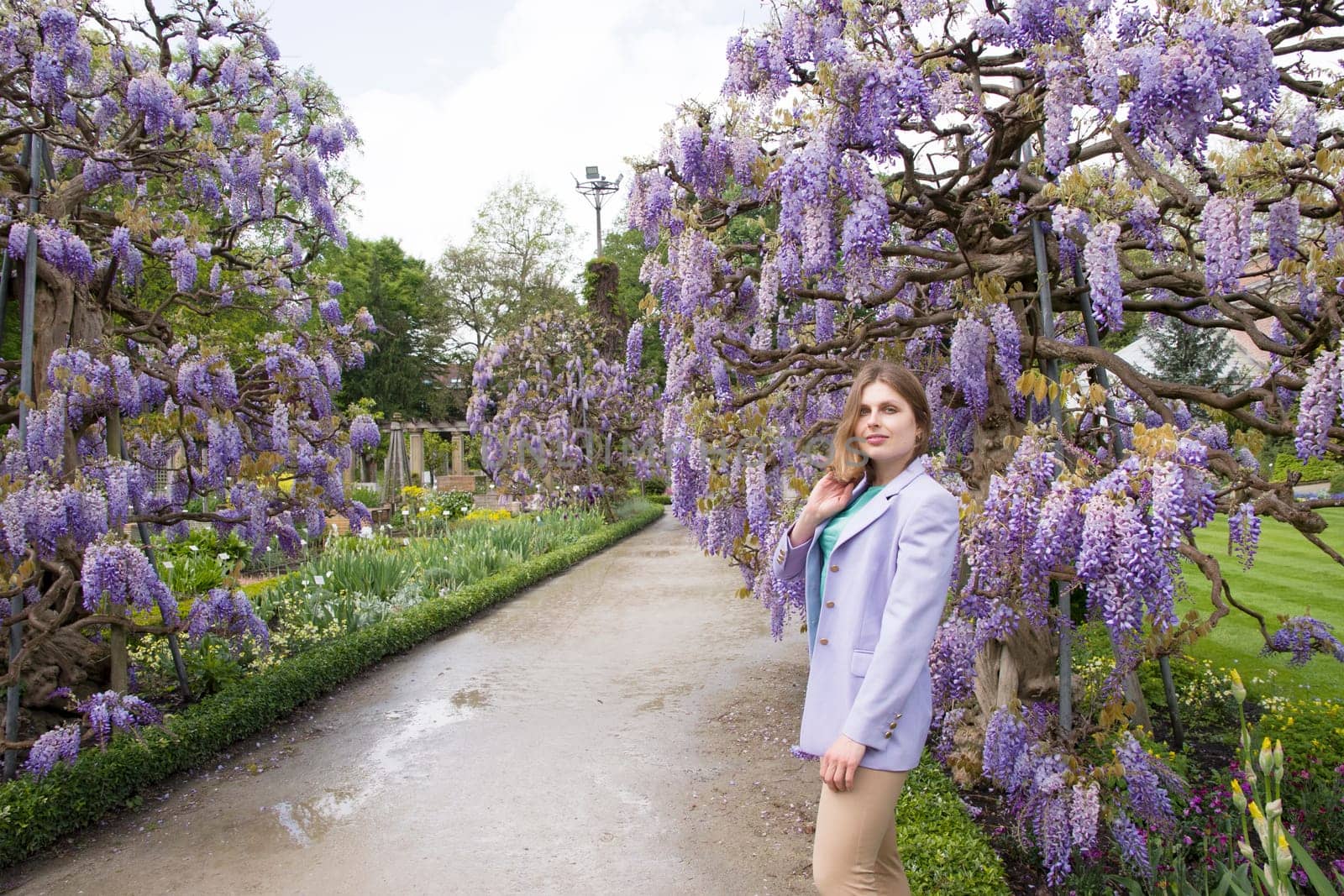 A young woman in a lilac jacket poses near the flowering wisteria in the Garden by KaterinaDalemans