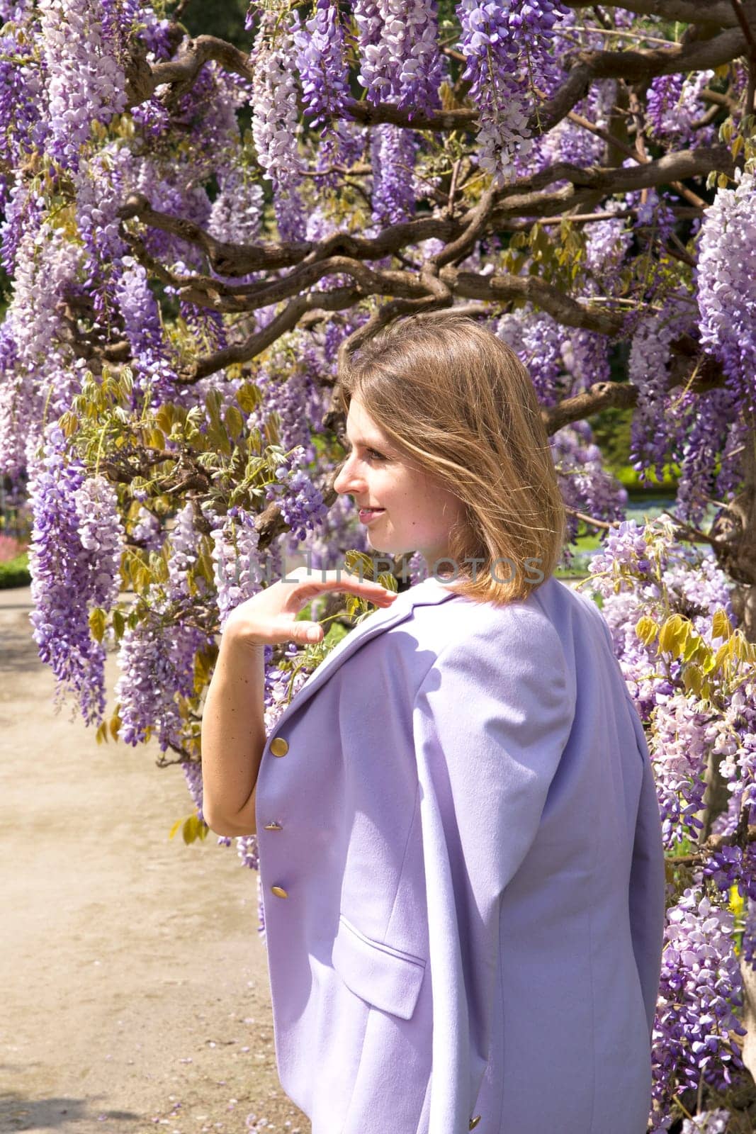 A young woman in a lilac jacket poses near the flowering wisteria in the Garden by KaterinaDalemans