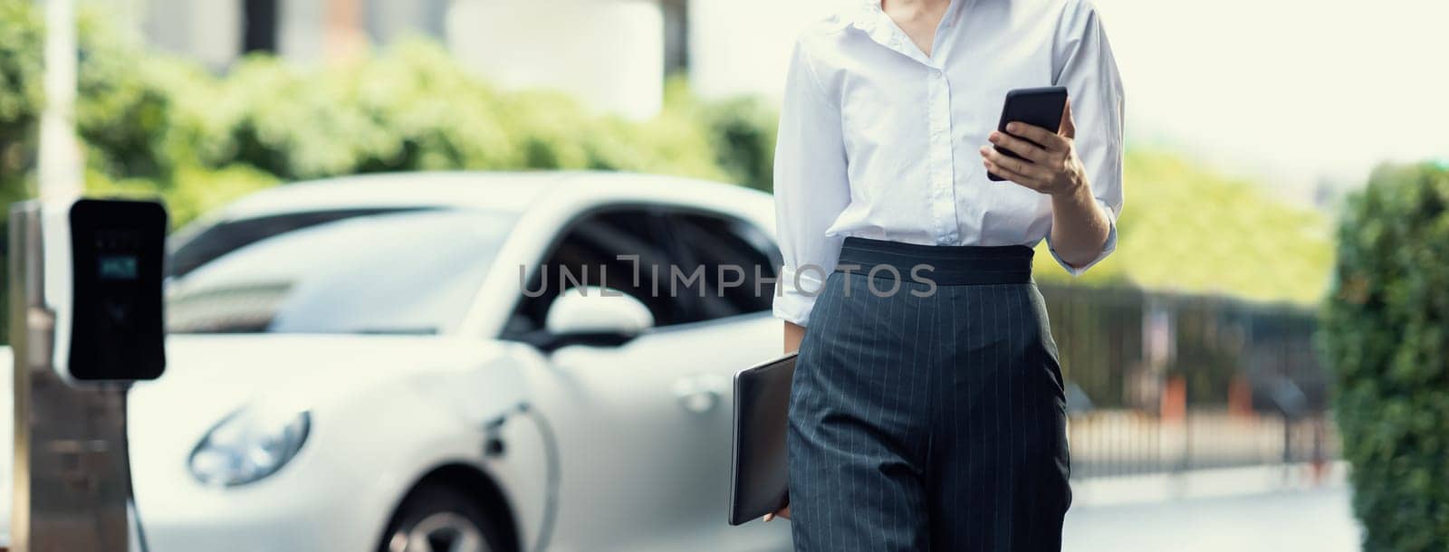 Closeup businesswoman using tablet, walking while recharging her electric vehicle with charging station at public car parking. Progressive lifestyle of technology and ecological concern by EV car