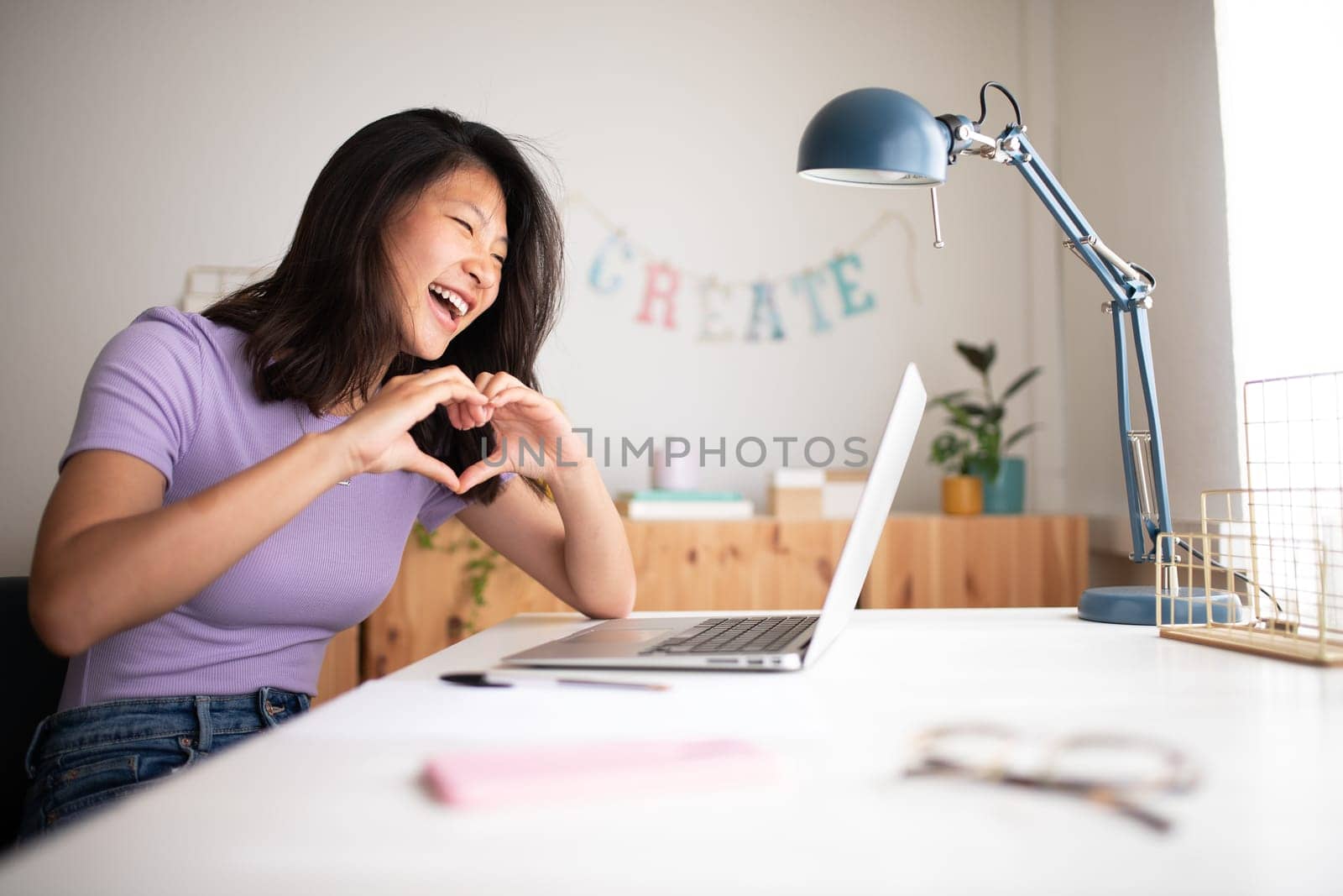 Happy teenage asian girl making heart shape with hands during online video call using laptop. Copy space. Love and technology concept.