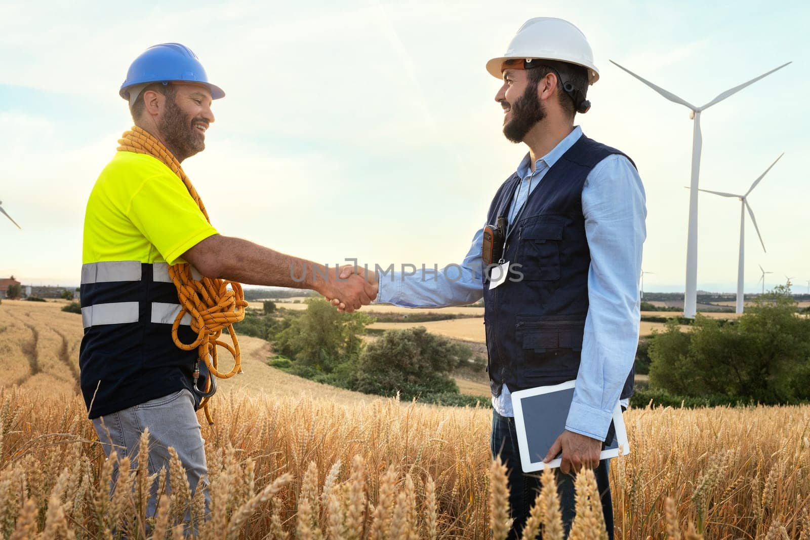Happy Male electric engineer and smiling maintenance worker shaking hands in wind turbine farm. Renewable energy.