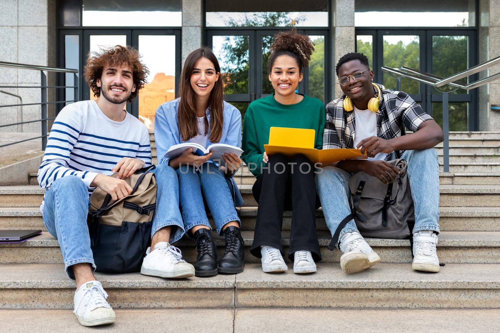 Portrait of happy and smiling multiracial group of college student friends looking at camera sitting on stairs. by Hoverstock
