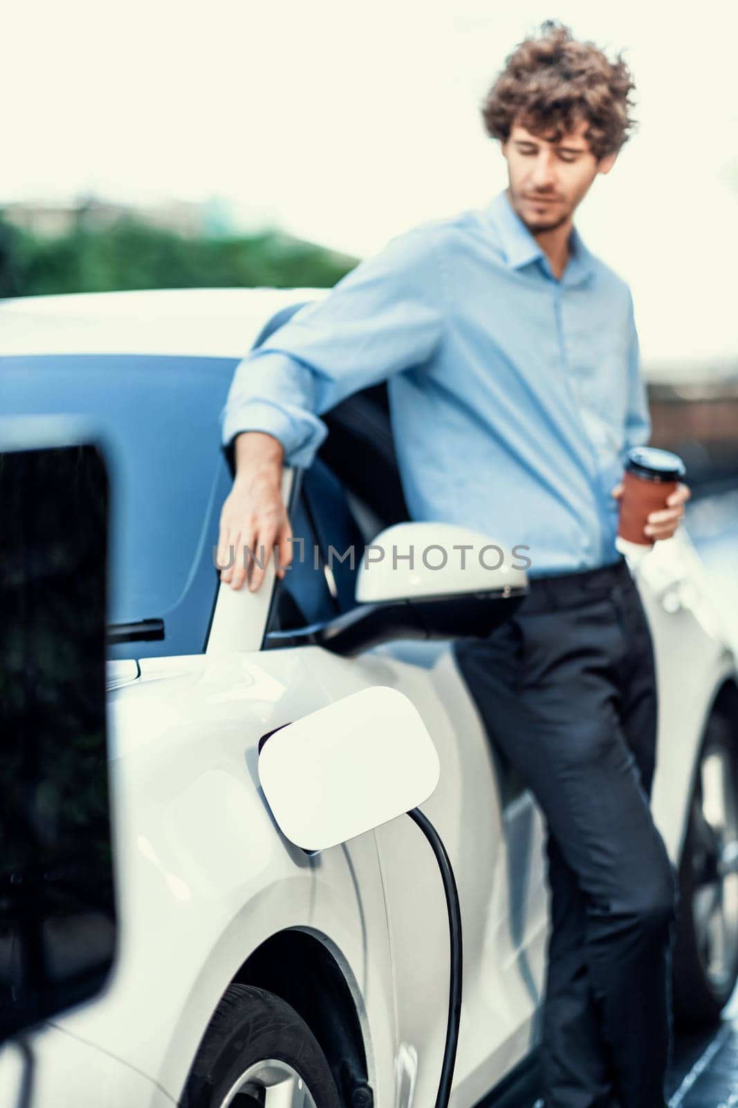 Progressive eco-friendly concept of focus parking EV car at public electric-powered charging station in city with blur background of businessman leaning on recharging-electric vehicle with coffee.