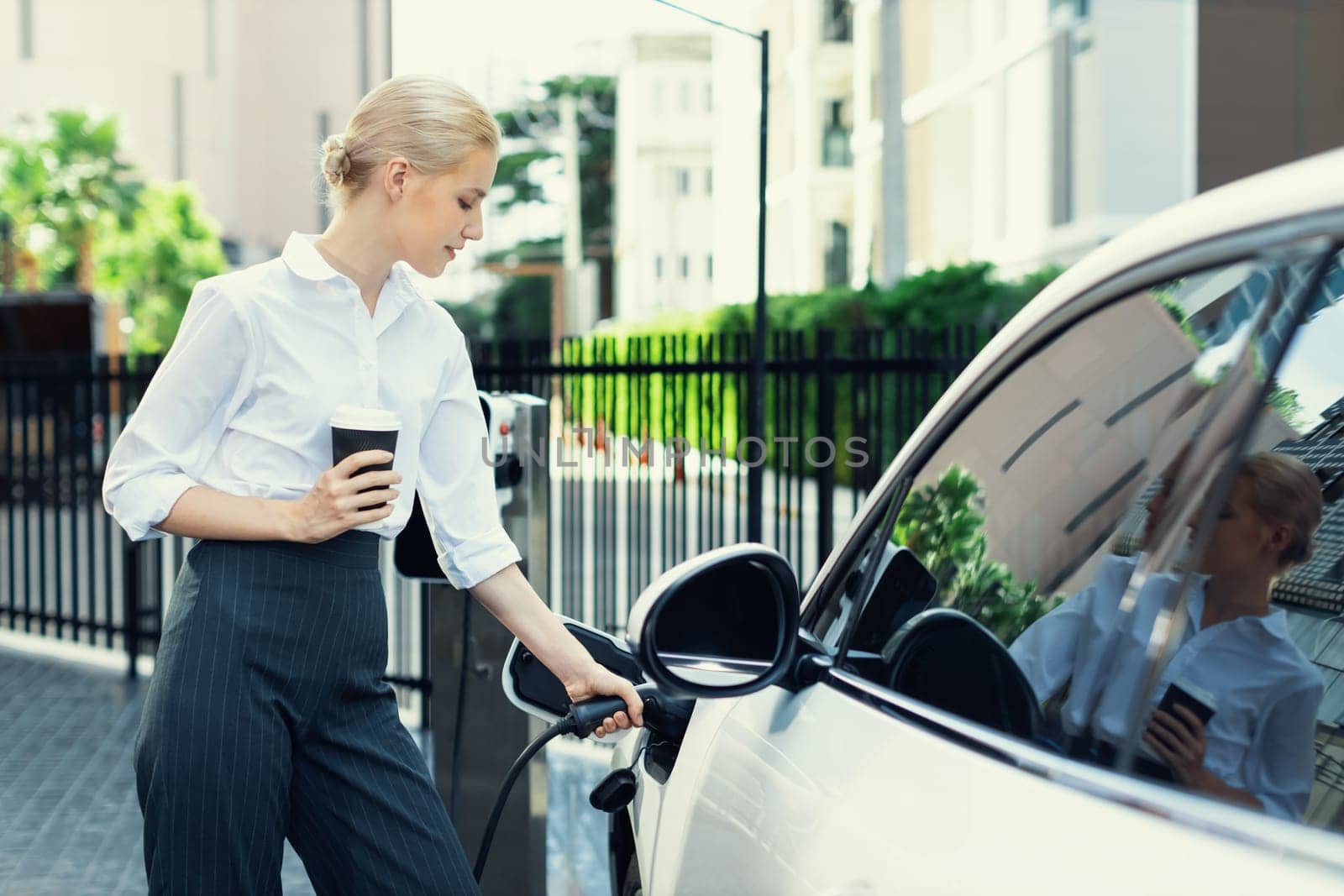 Businesswoman drinking coffee, leaning on electric vehicle recharging at public charging station with residential apartment condos building in background as progressive lifestyle by eco-friendly car.