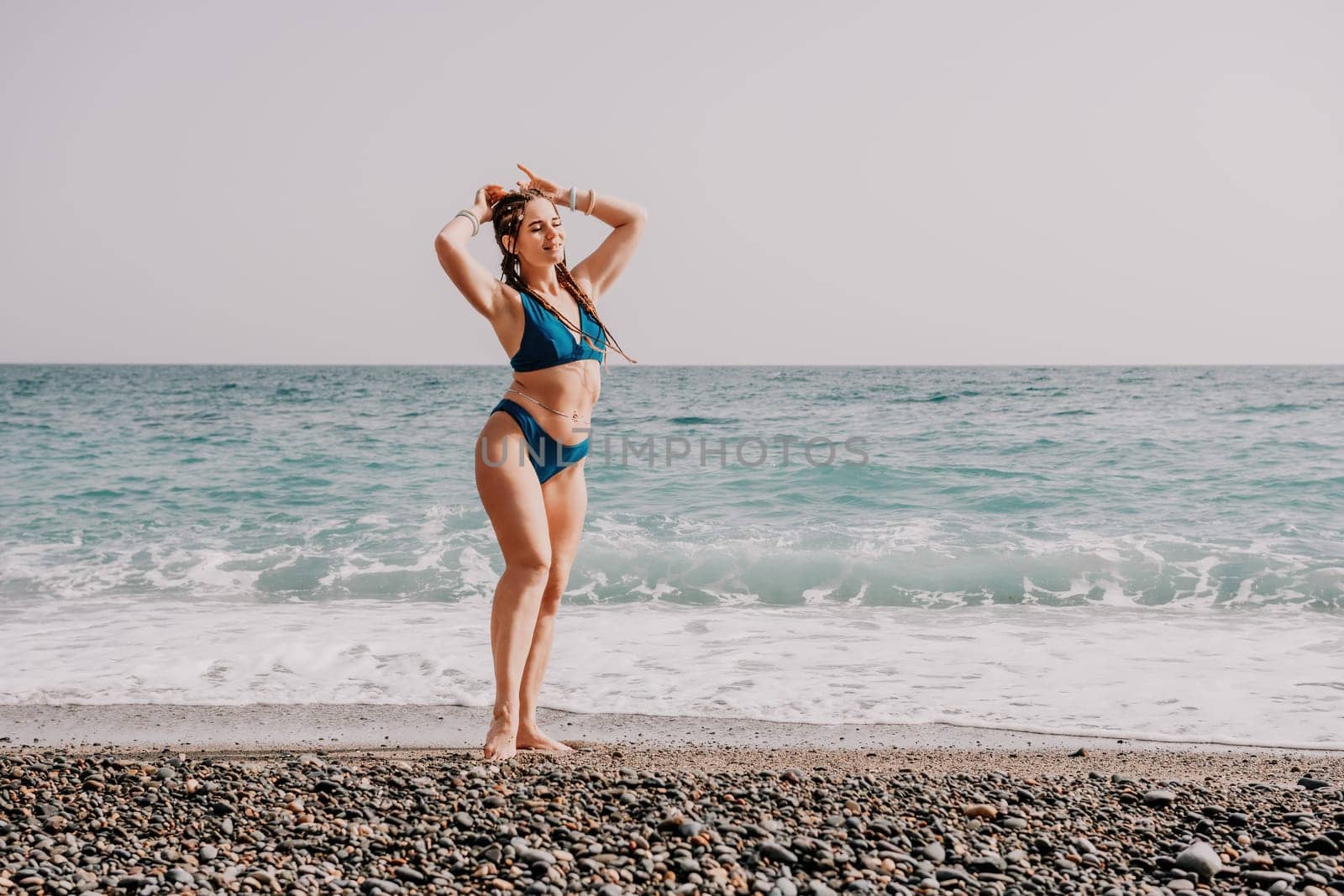 Beach vacation. Hot beautiful woman in sunhat and bikini standing with her arms raised to her head enjoying looking view of beach ocean on hot summer day.