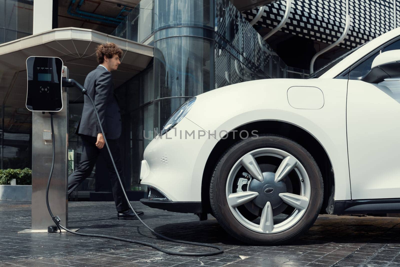 Below view of progressive businessman with electric car recharging at public charging station at modern city residential building. Eco friendly rechargeable EV car powered by alternative clean energy.