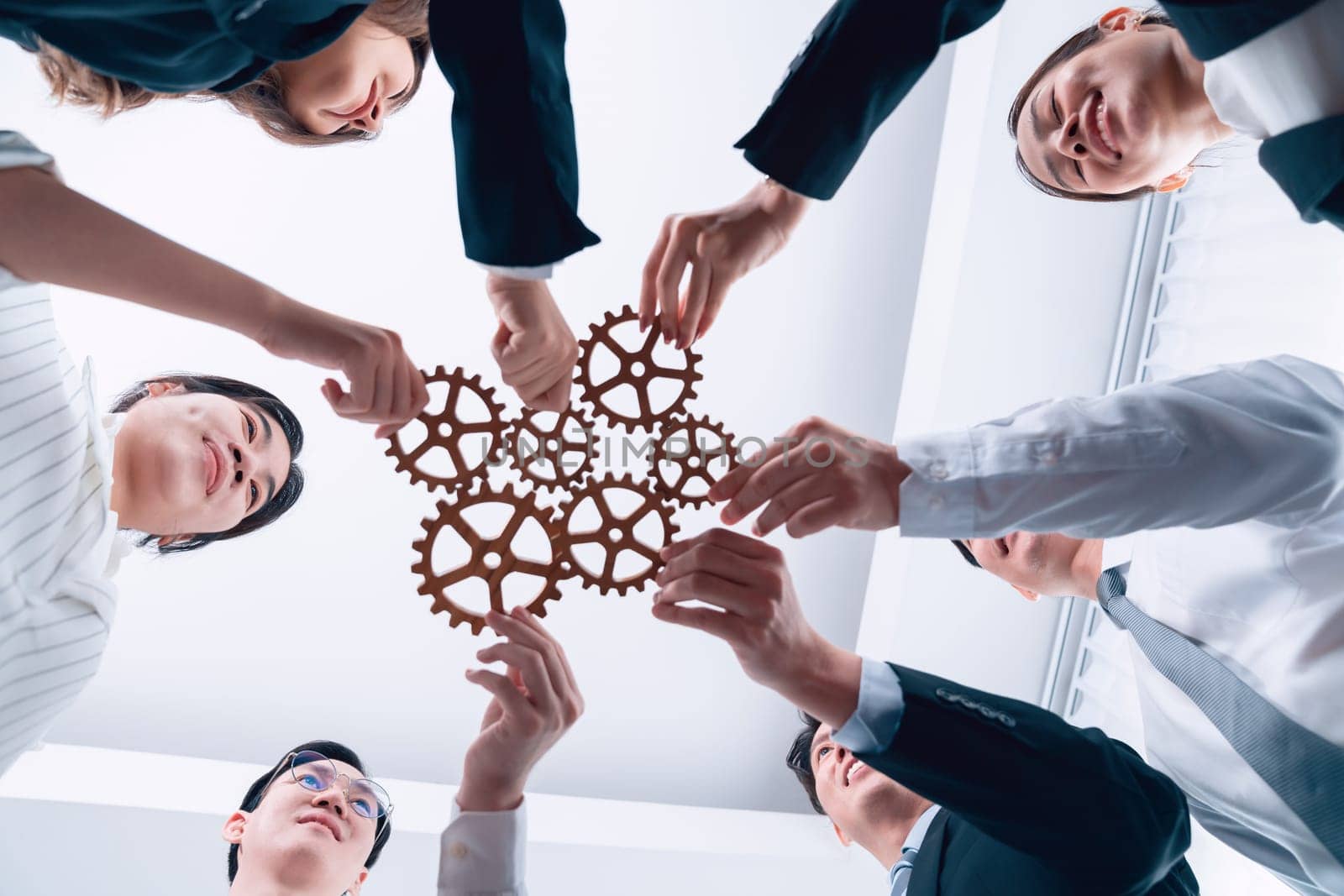 Hand holding wooden gear by businesspeople wearing suit for harmony synergy in office workplace. Bottom view office worker hand make chain of gear into collective unity form.