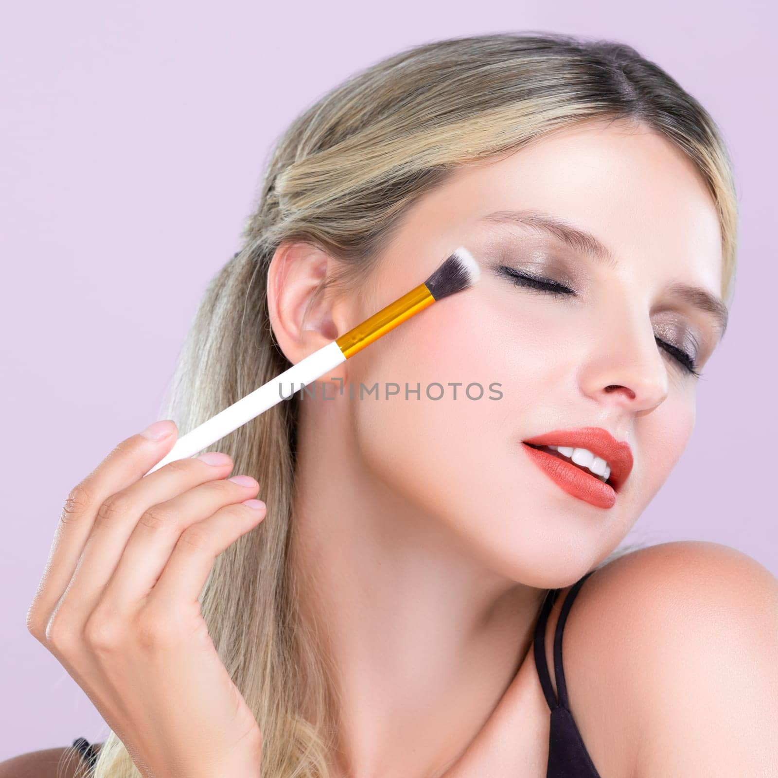Closeup beautiful girl with flawless applying alluring eye shadow makeup with eyeliner brush. Cosmetic facial painting process on lovely young woman with perfect clean skin on pink isolated background
