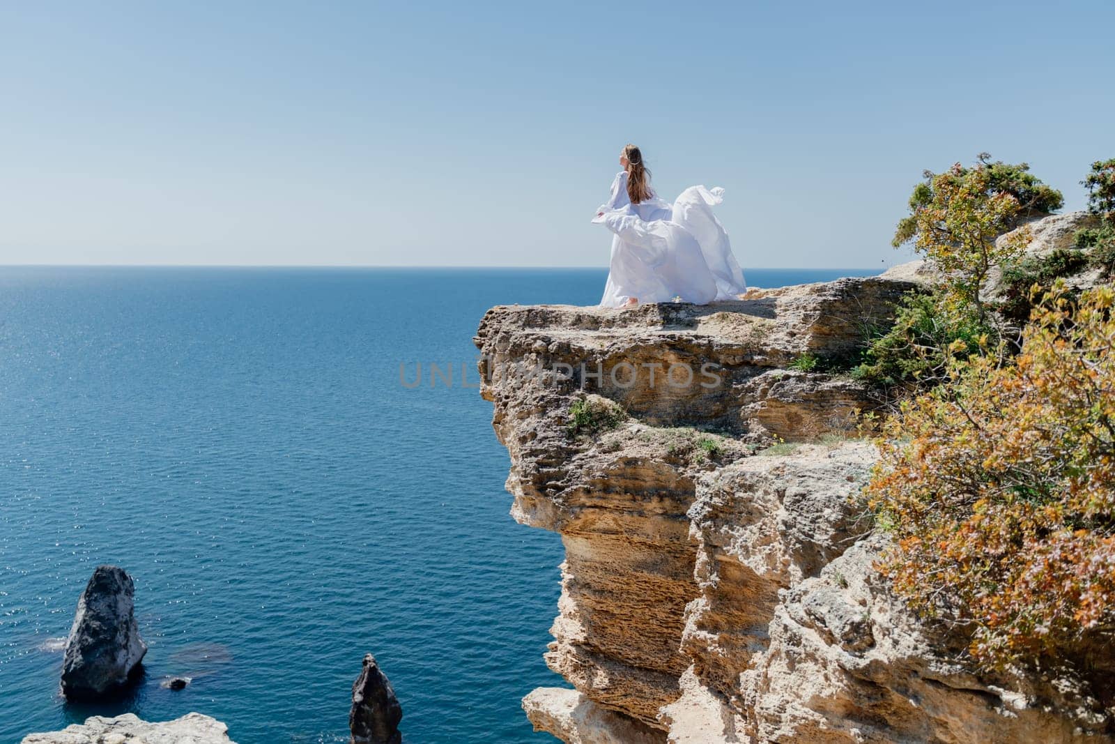 Woman in a white dress on the sea. Side view Young beautiful sensual woman in white long dress posing on a rock high above the sea at sunset. Girl in nature against the blue sky by Matiunina