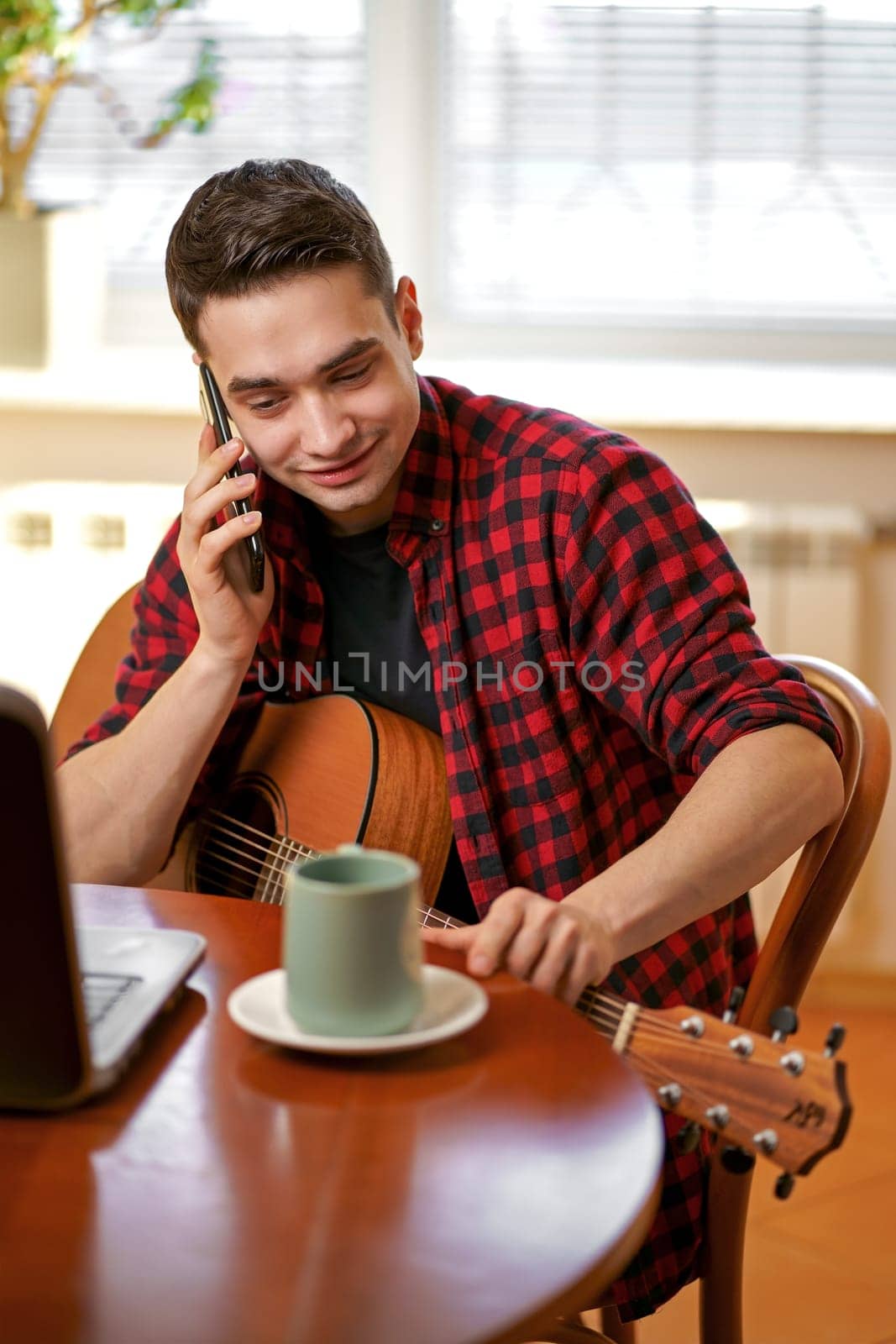 Relaxed young man playing guitar at home and using laptop, drinking tea, laughing and talking on the phone by aprilphoto
