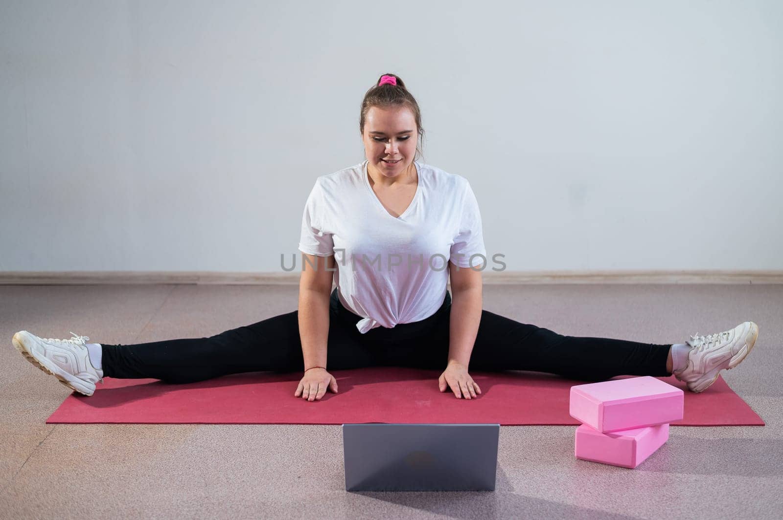 Young caucasian fat woman doing bends on a sports mat and watching a training video on a laptop. A chubby girl stretches the split remotely using video communication.