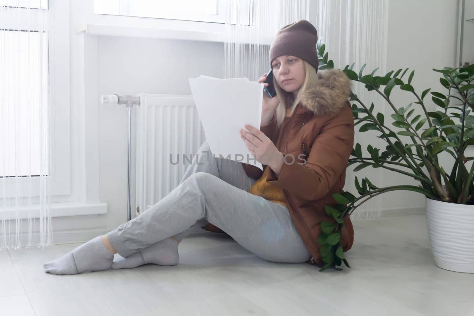 A woman warmly dressed in a brown jacket and hat is sitting near the heater with bills in her hands. The concept of a crisis and a large set for utilities