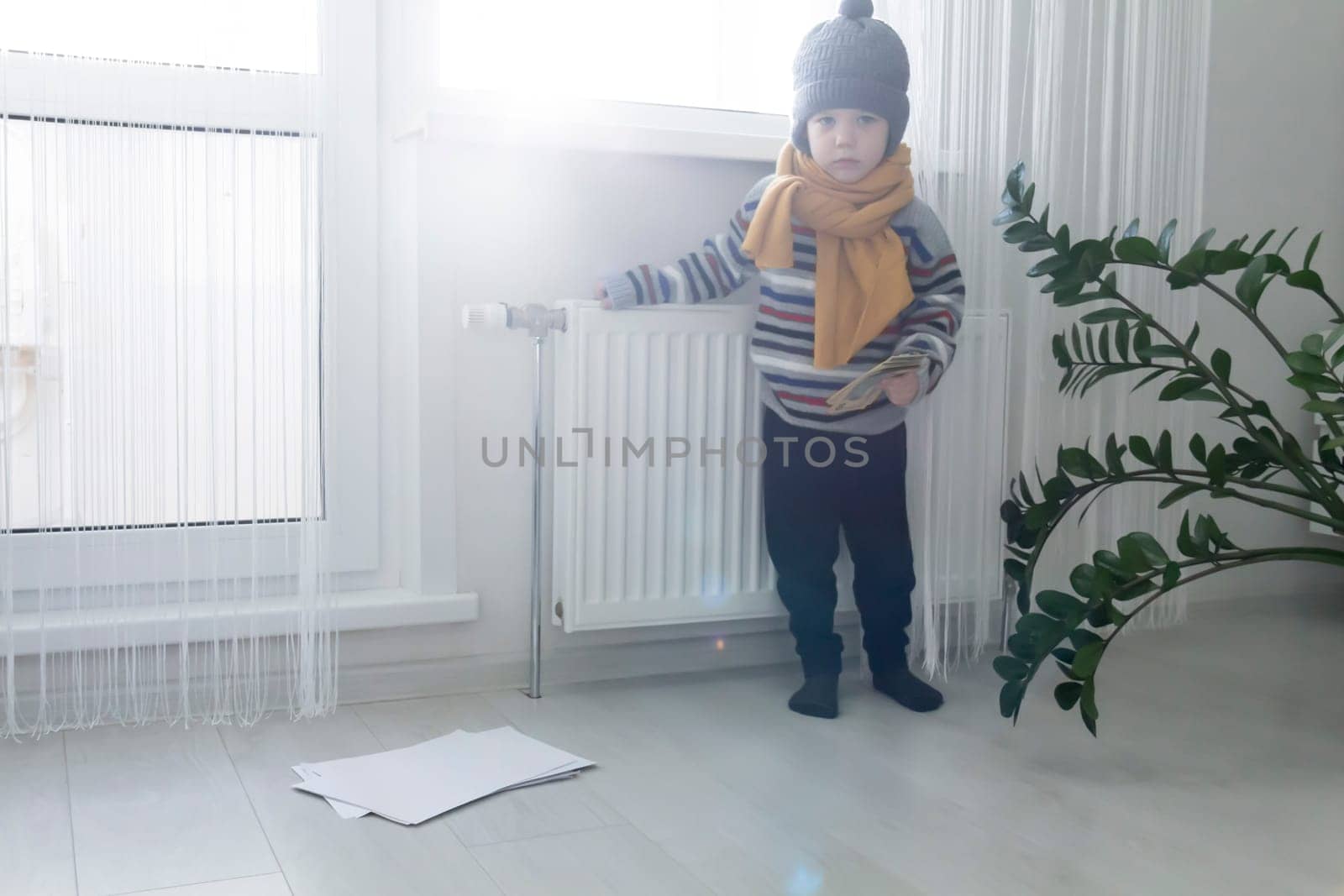 The child is warmly dressed in a sweater and a hat, standing and holding on to the radiator with money and bills. The concept of the economic crisis and the lack of heat and heating in homes