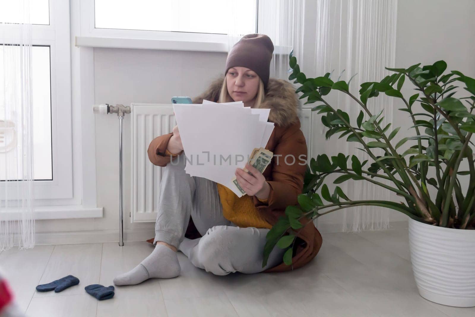 A woman warmly dressed in a brown jacket and hat is sitting near a heating appliance with bills in her hands, money and a phone. The concept of a crisis and a large set for utilities by Alla_Yurtayeva