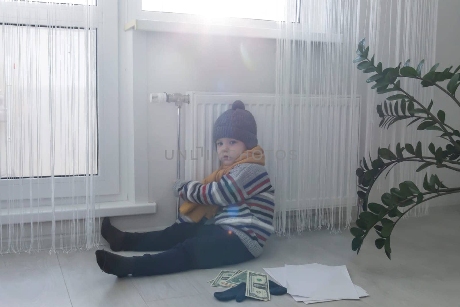 The child is warmly dressed in a sweater and a hat, sitting and holding on to the radiator. The concept of the economic crisis and the lack of heat and heating in homes