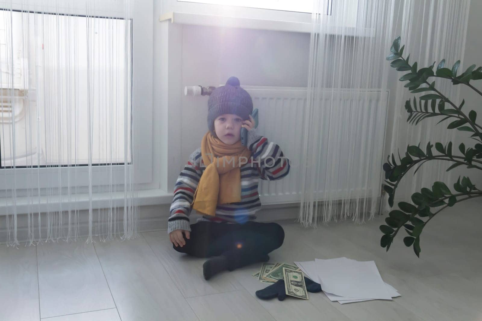 The child is warmly dressed in a sweater and a hat, sitting near the heater with bills in her hands, money and a phone. The concept of a crisis and a large set for utilities. by Alla_Yurtayeva
