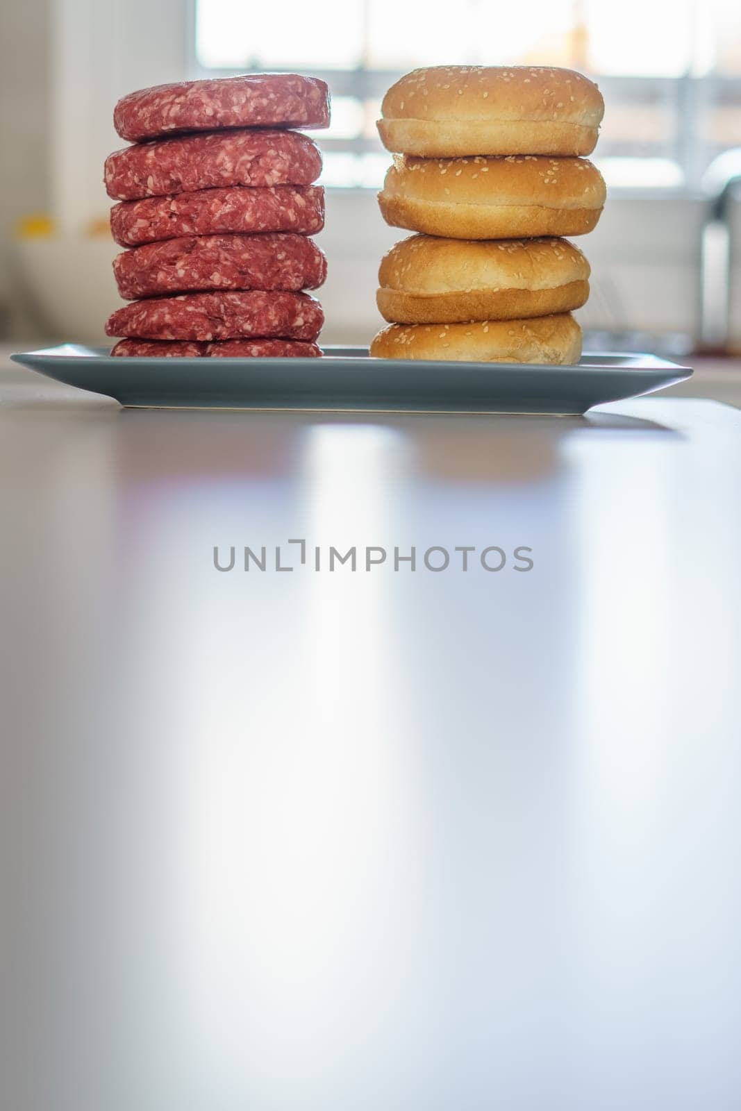 stack of raw beef burgers with their buns on a mirrored white plate